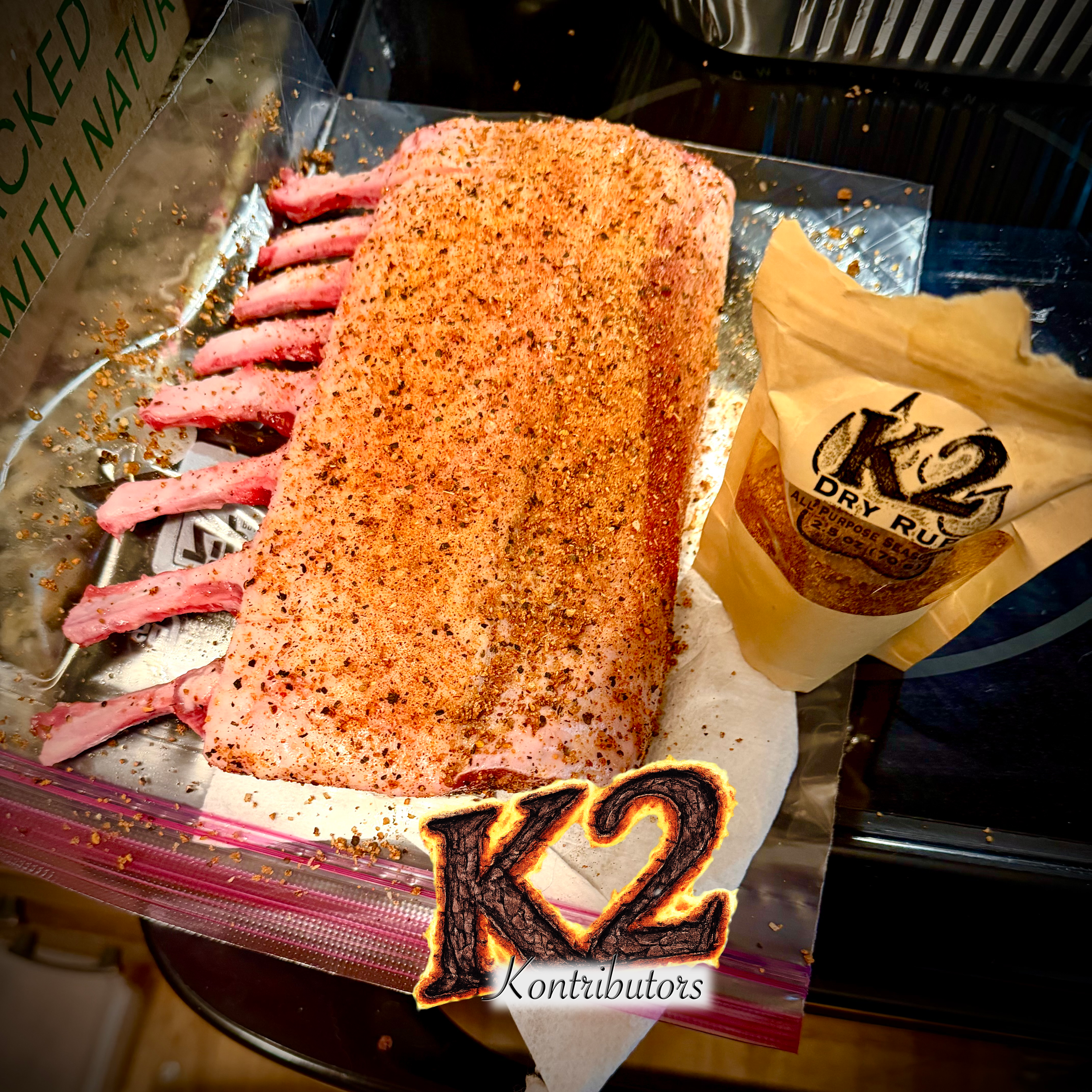 A prepared rack of lamb, seasoned with k2BBQ spices, resting on a foil-lined tray. Part of a packaging bag is visible nearby.