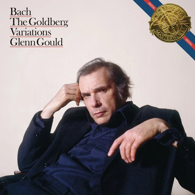 Album cover of Bach, The Goldberg Variations by Glenn Gould featuring a portrait of Glenn Gould sitting in a dark suit with a serious expression, resting his head on his hand, against a plain background, with a gold record badge in the upper right corner.