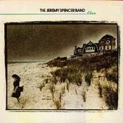 A person crouching on sandy dunes near a house by the beach, with grass and dunes in the foreground and the house in the background.