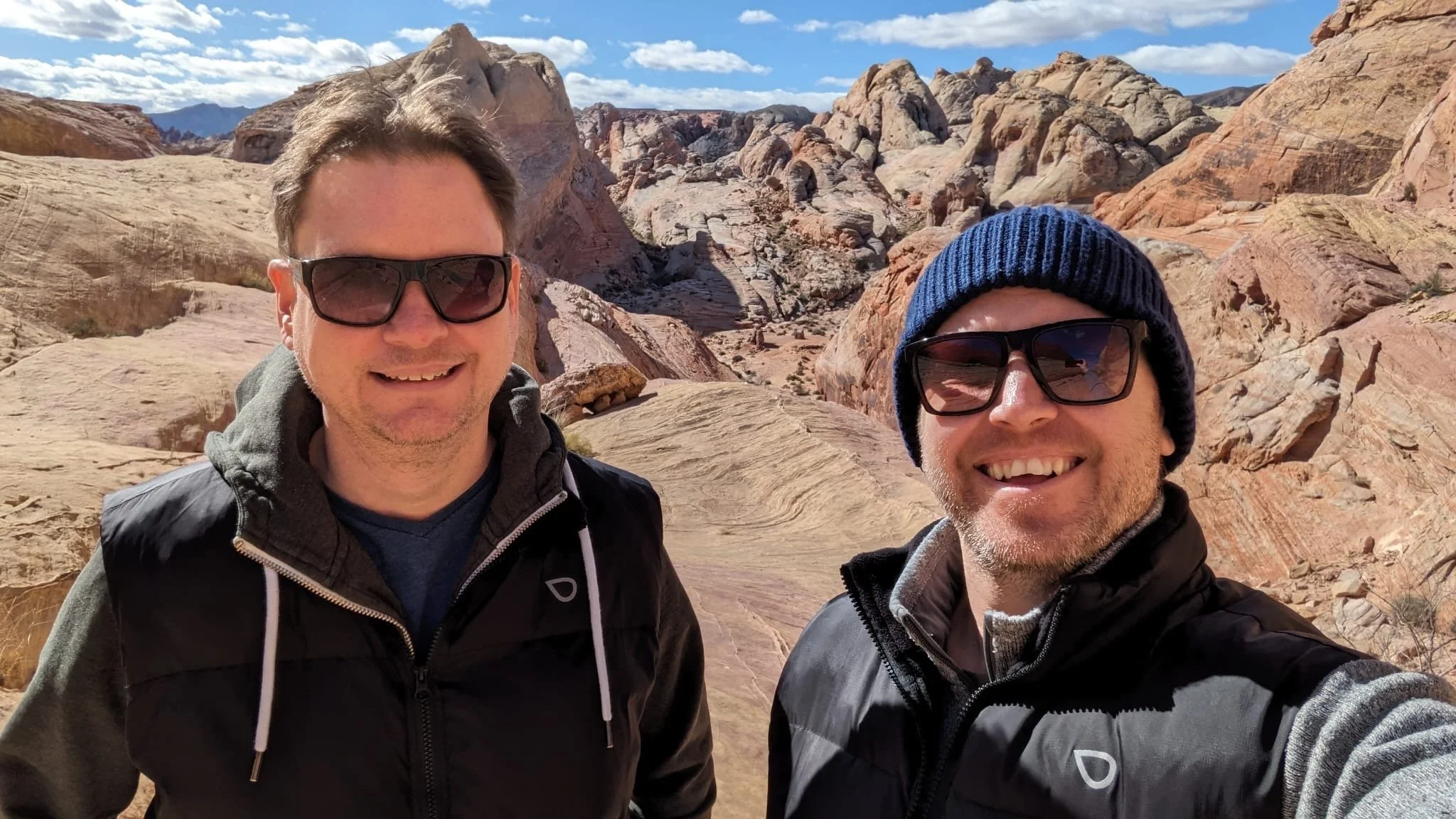 Two men smiling in a desert landscape with large rock formations and a partly cloudy sky.