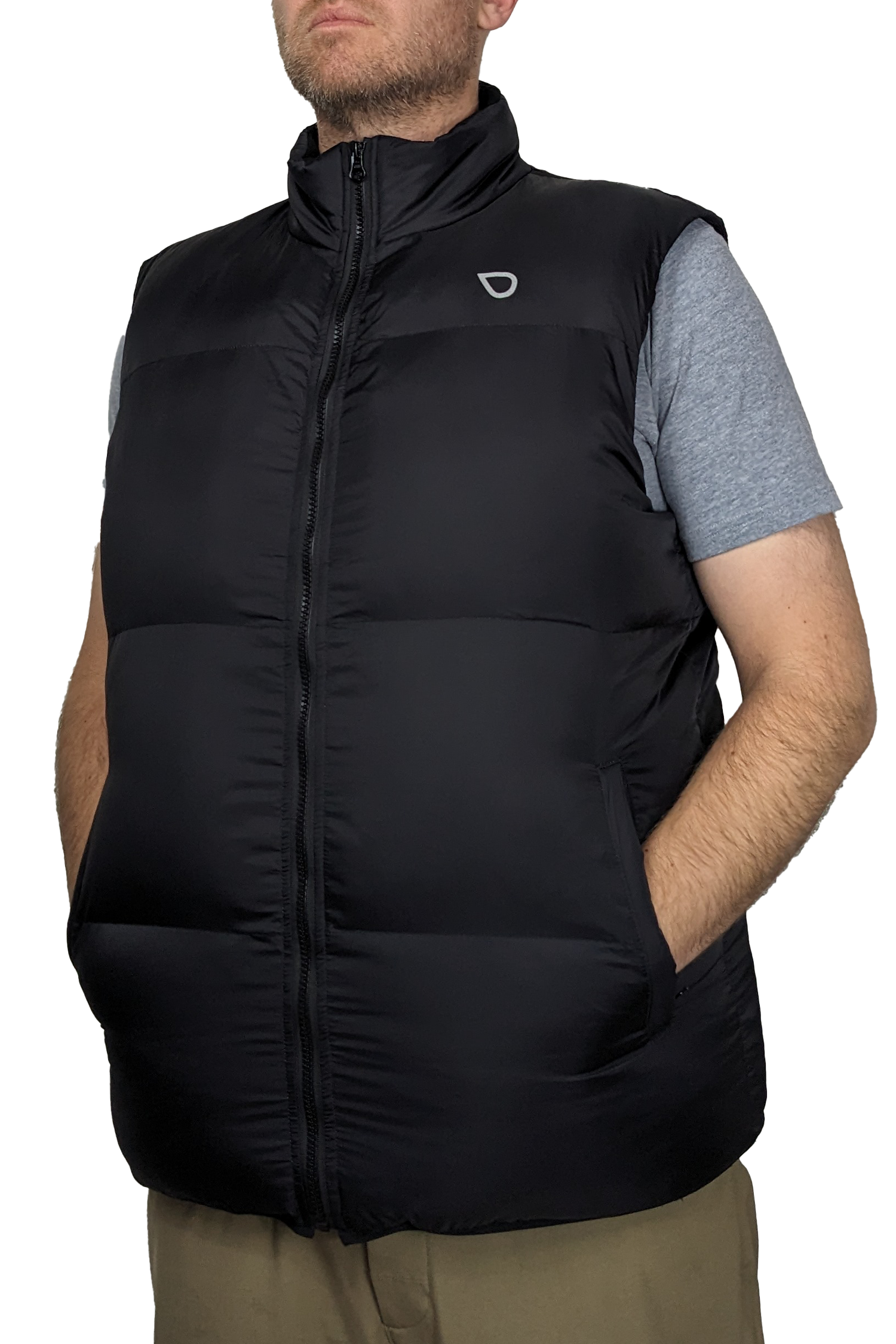 A man wearing a black puffer vest over a gray T-shirt, with hands in his pockets, standing against a plain background. The jacket contains clothing avoiding the need for a cabin bag. This avoids the airline fees charged by Ryanair, Easyjet, Wizzair