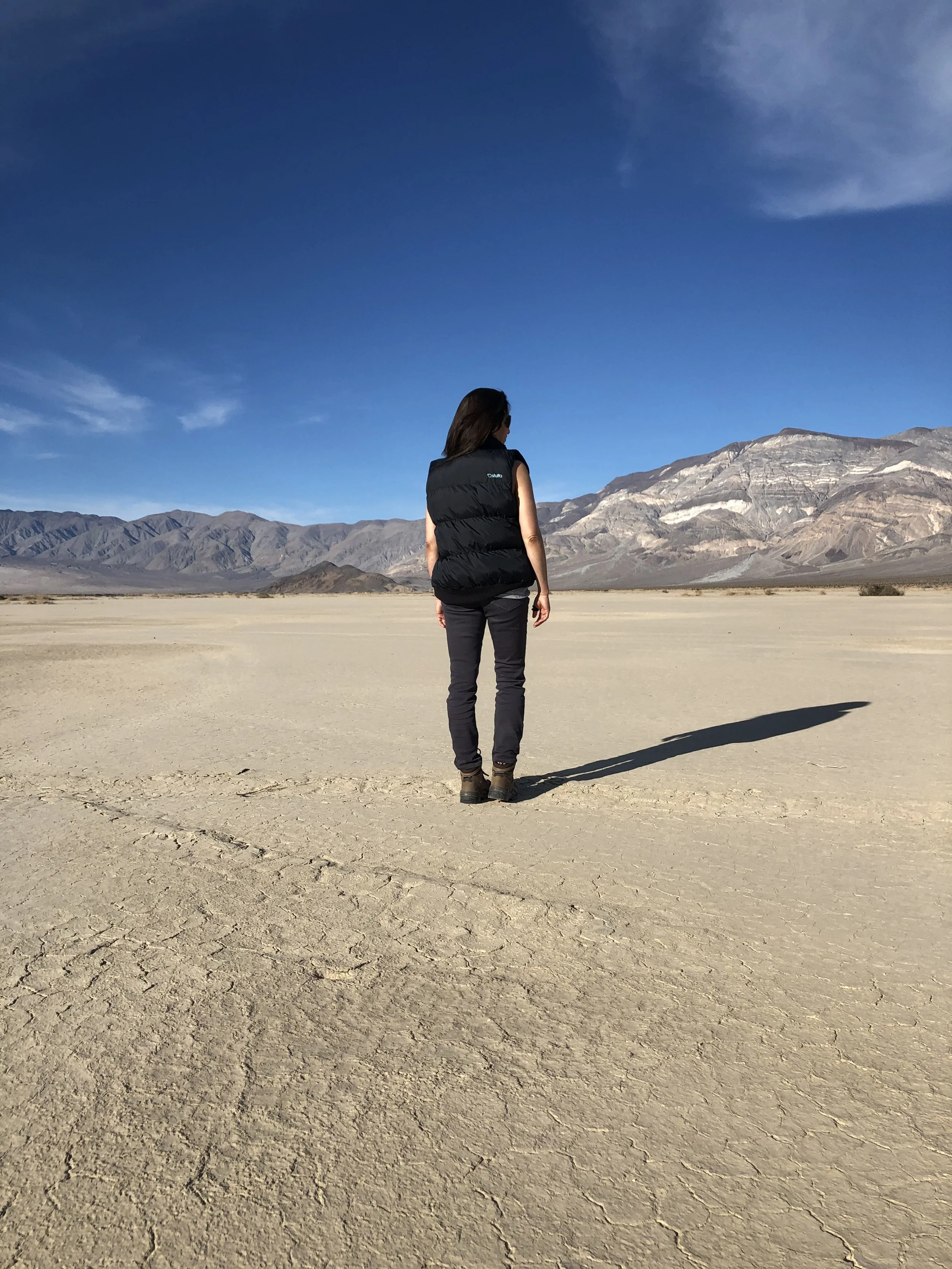 A beautiful woman standing alone in a vast, dry desert landscape with mountains in the background and a blue sky overhead.