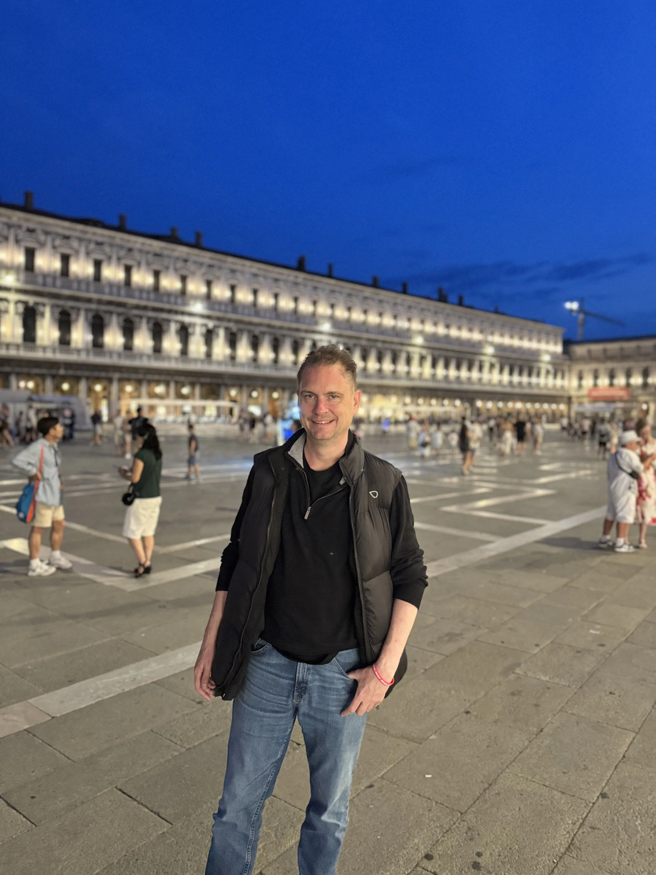 A man in a black jacket and blue jeans standing in a busy square at night, with historical buildings illuminated in the background, and a clear blue evening sky. His jacket contains 8 days of clothing inside the hidden pockets of the jacket.