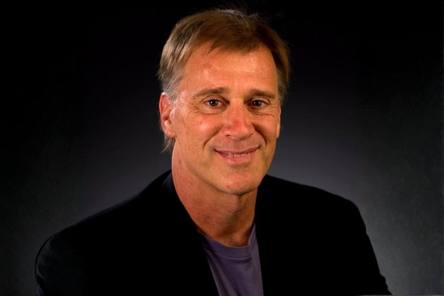 Portrait of a middle-aged man with short light brown hair, smiling, wearing a dark blazer and a gray shirt, against a dark background.