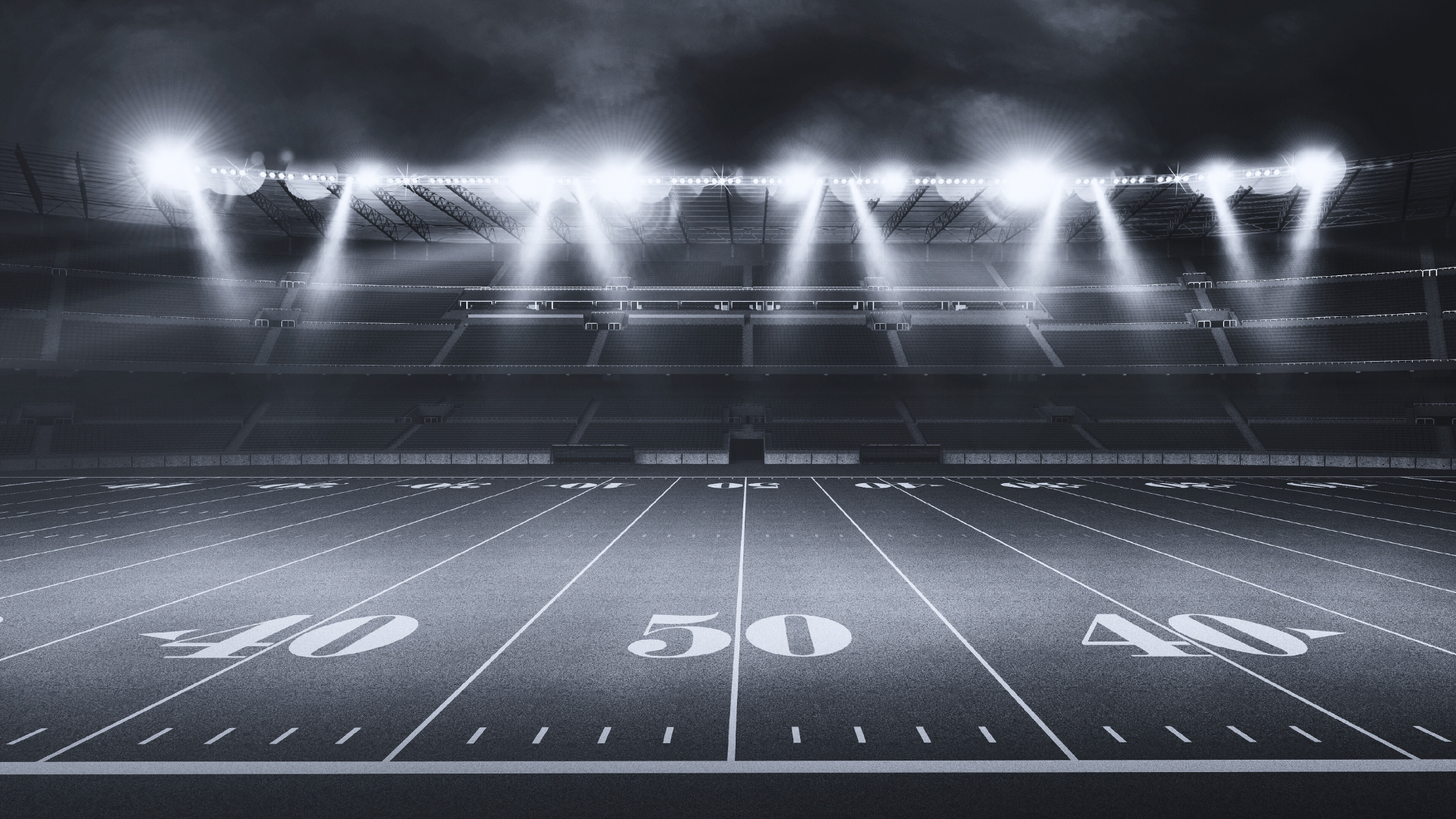 Empty football stadium at night with bright floodlights illuminating the field and dark cloudy sky overhead.