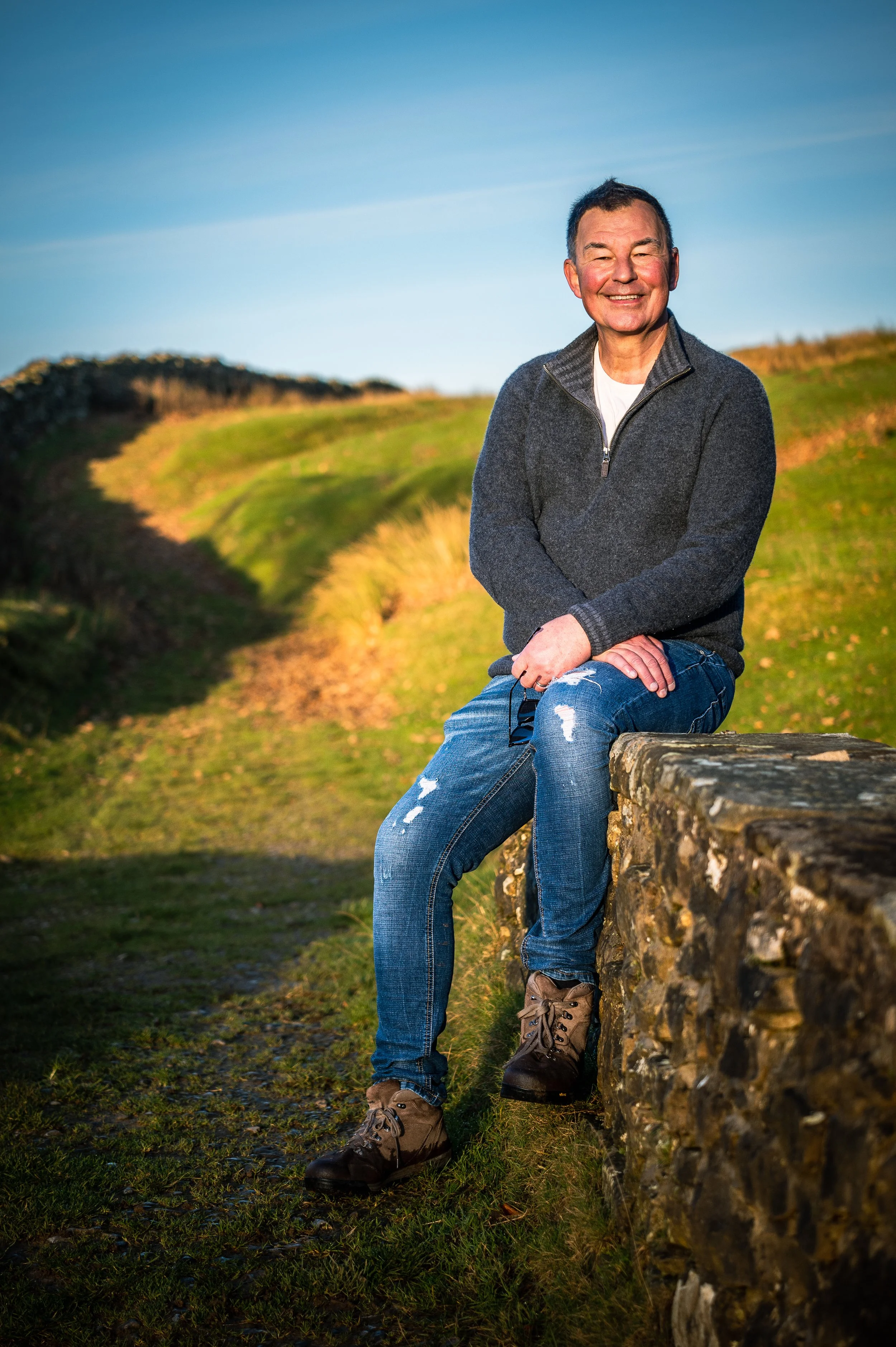 A smiling man sitting on a stone wall outdoors with green grass and hills behind him, under a clear blue sky.
