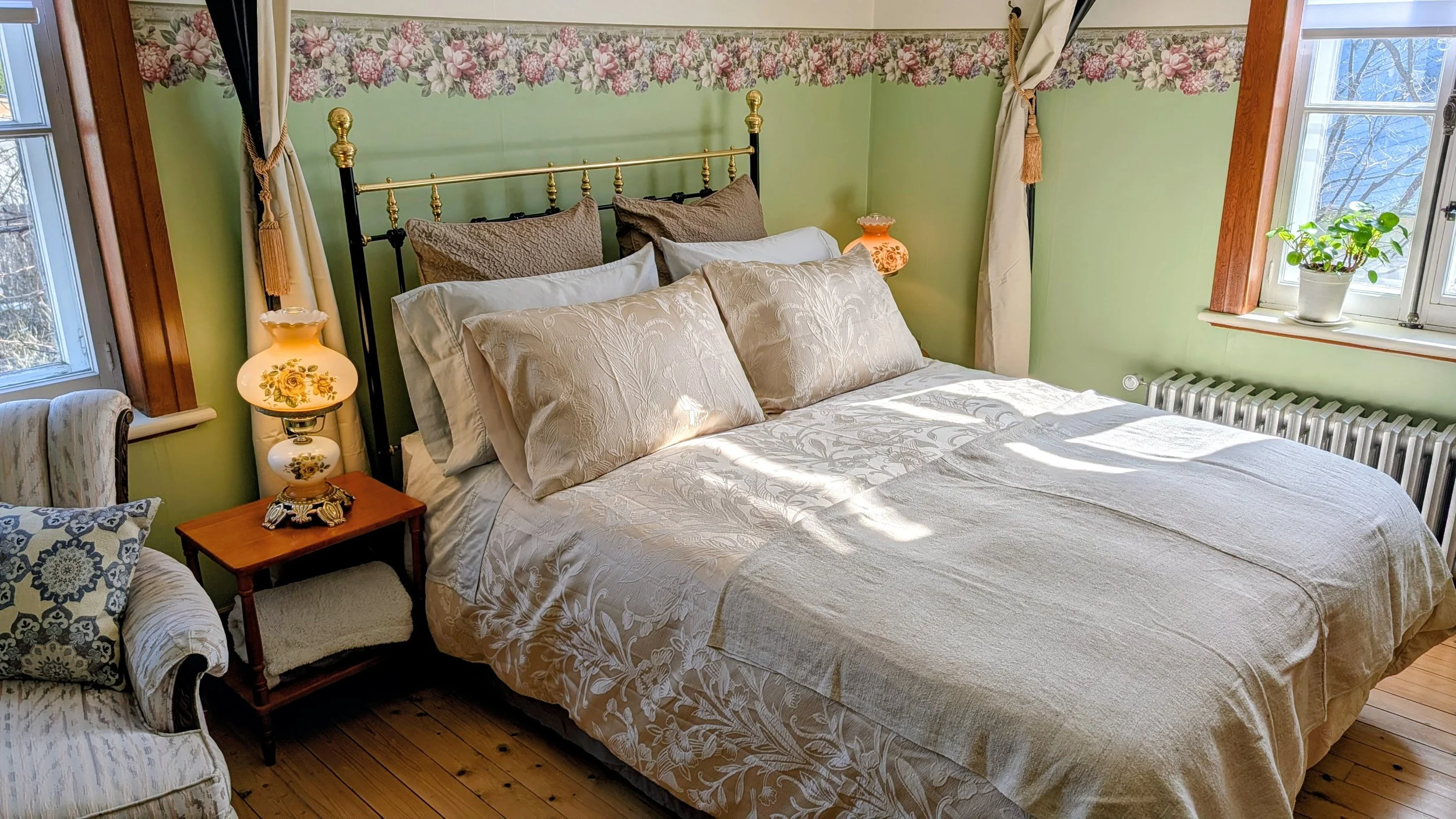 Chambre avec lit double, coussins en tissu beige et bronze, lampes à chaque côté, fenêtres donnant sur un jardin avec neige.