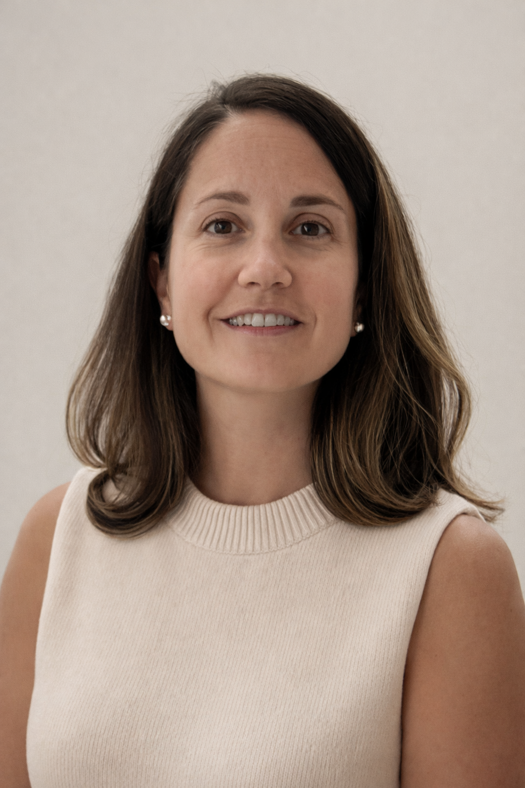 Portrait of a woman with shoulder-length brown hair, wearing a sleeveless beige top and pearl earrings, smiling softly against a plain light-colored background.