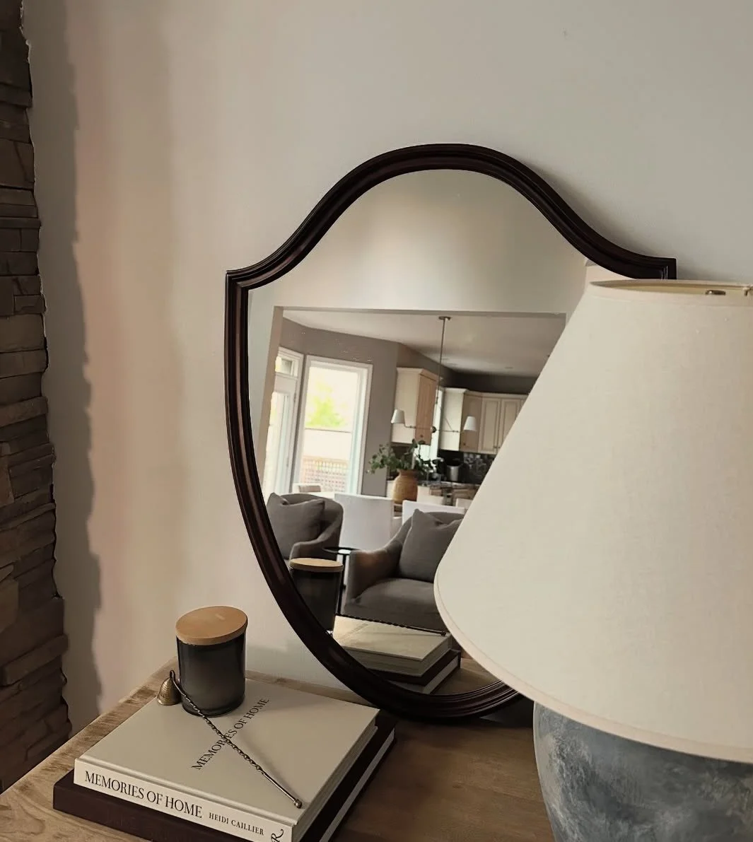 A heart-shaped mirror on a wall, reflecting a living room and kitchen area with large windows, plants, and furniture, with a lamp and books in the foreground.