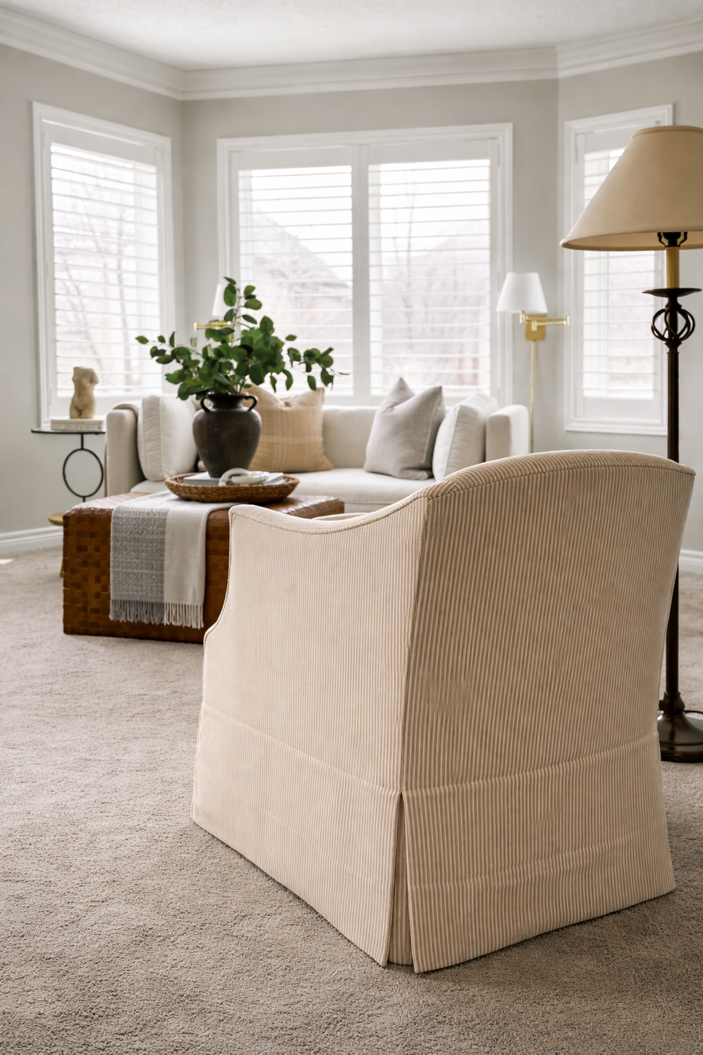 Living room with beige armchair in the foreground, cream sofa with cushions, coffee table with green plant in a dark vase, and large windows with white blinds, creating a bright, cozy space.