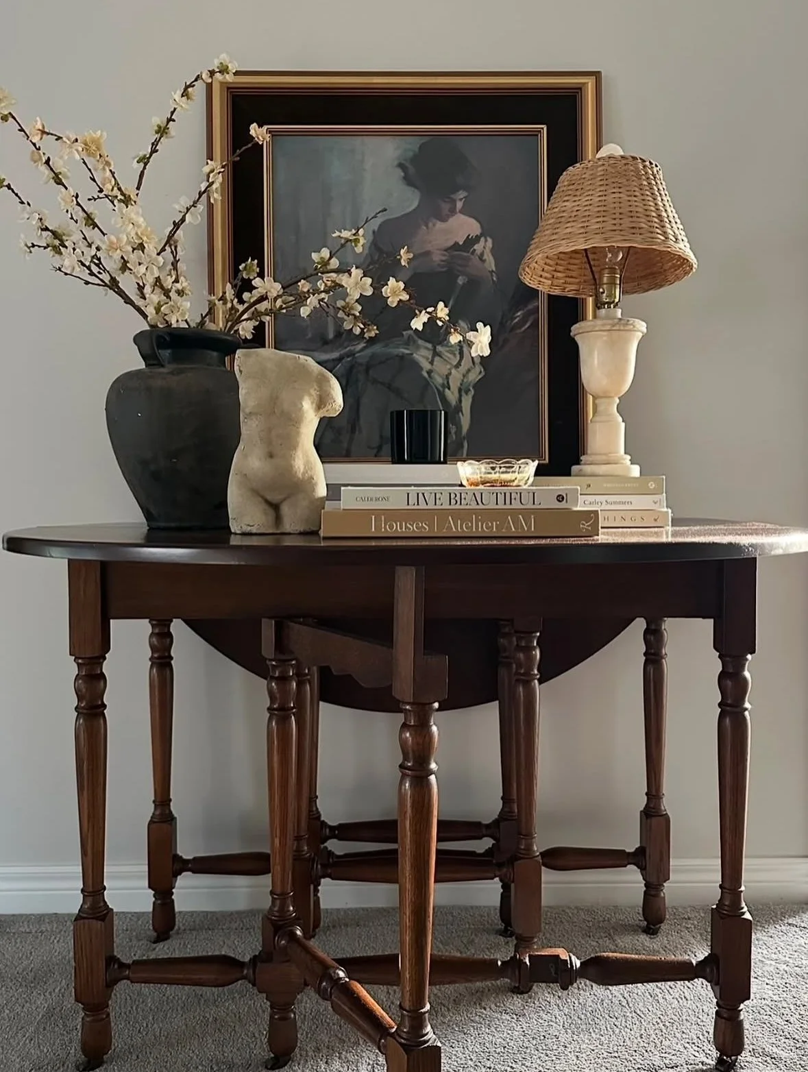 Decorative interior scene with a wooden table, topped with books, vases, and a lamp, against a wall with framed artwork and a decorative flower arrangement.