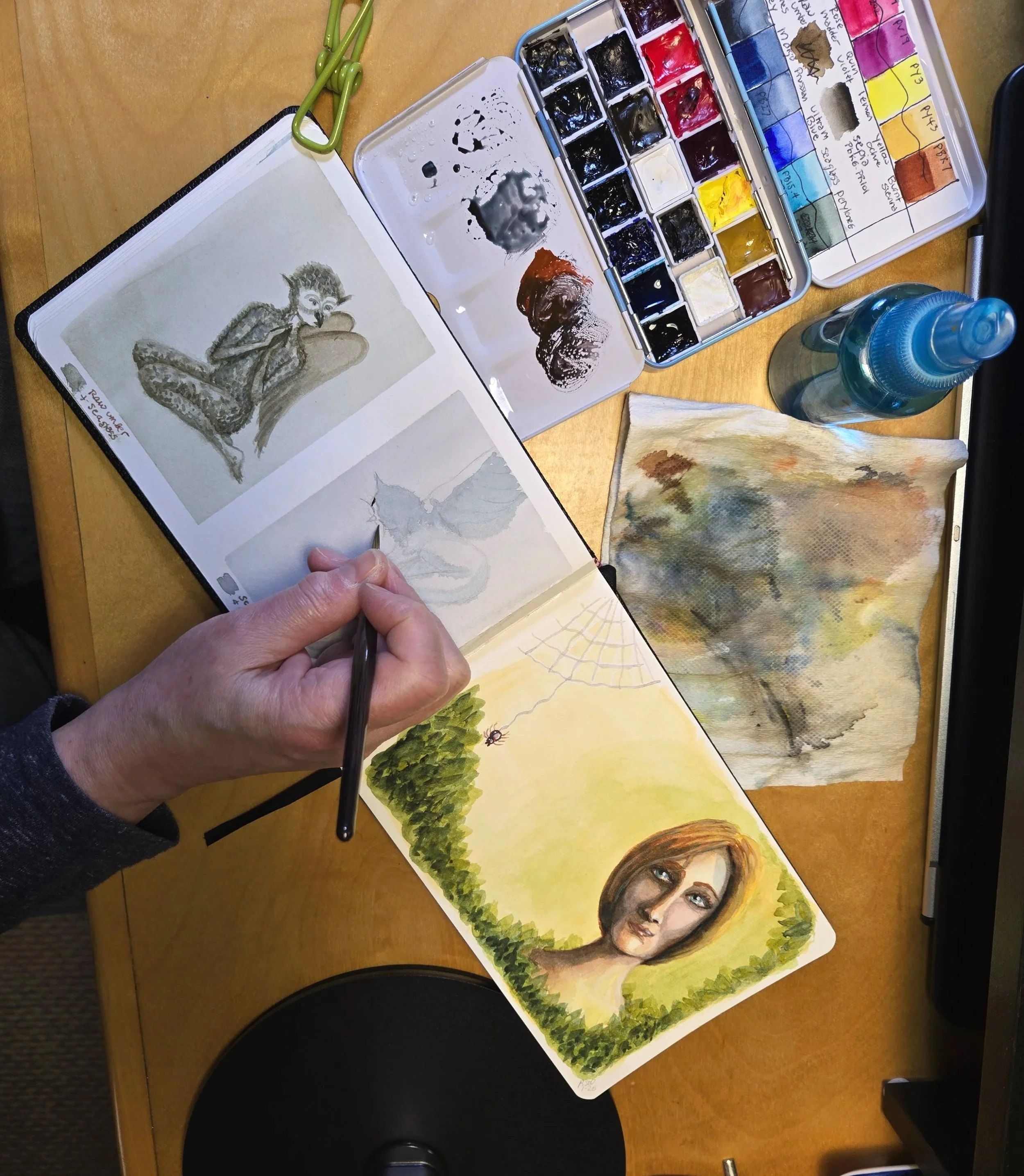 Open sketchbook with a grayscale drawing of a dragon or wyvern and a watercolor painting of a woman’s face with a green landscape background, surrounded by watercolor paints, brushes, a water bottle, and a piece of watercolor paper on a wooden desk.