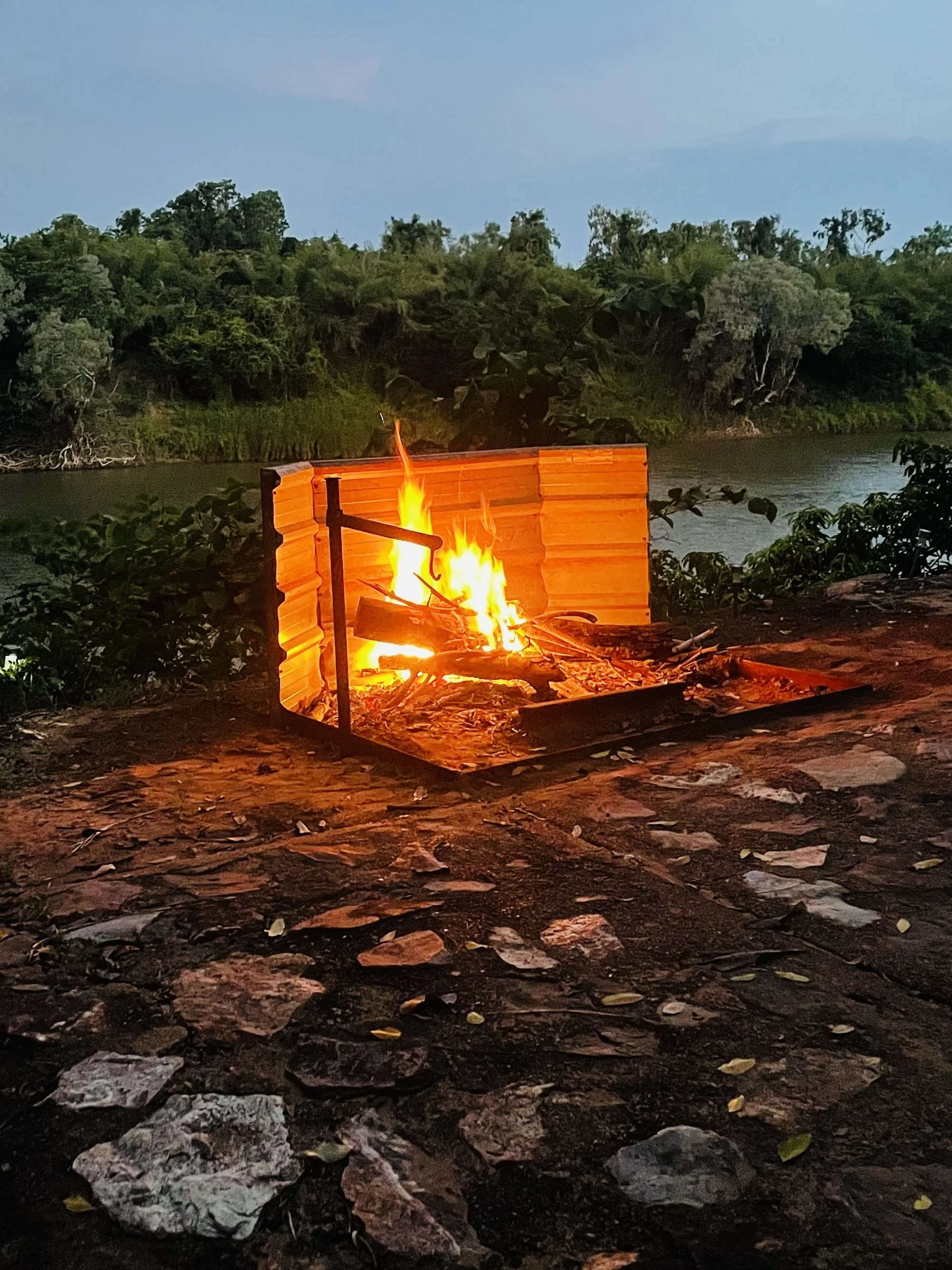 A small campfire burns within a metal fire pit on a rocky ground near a river, with trees and woods in the background and a cloudy sky.