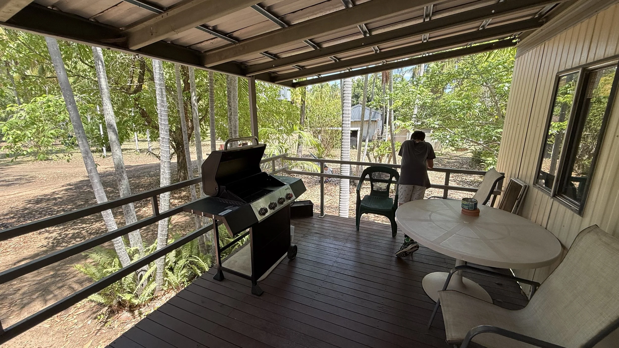 A person standing on a covered porch, looking out at a wooded backyard with trees and a shed, with a grill and outdoor furniture present.
