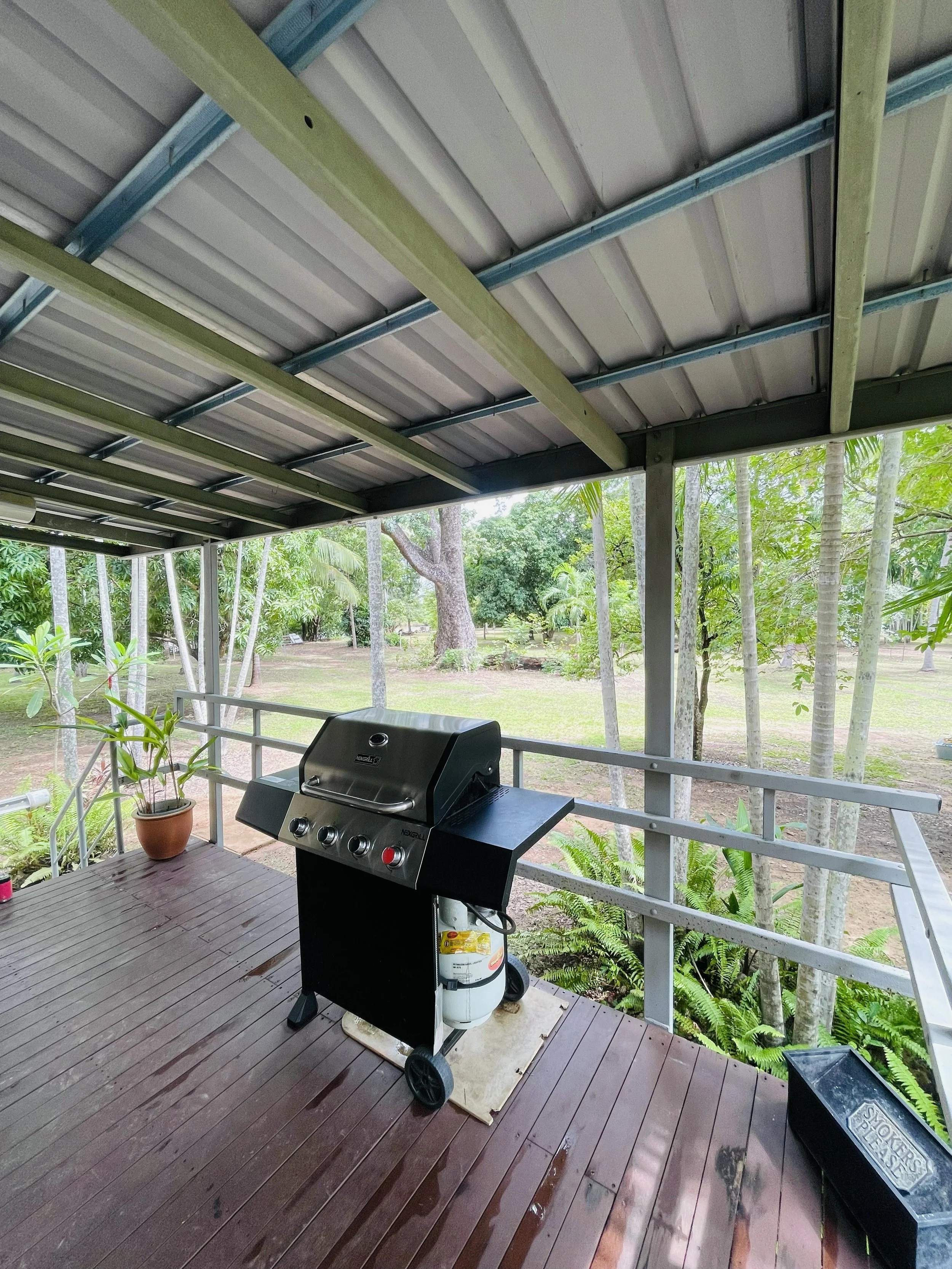 Outdoor patio with a propane gas grill on a wooden deck, surrounded by green trees and plants, under a metal roof canopy.