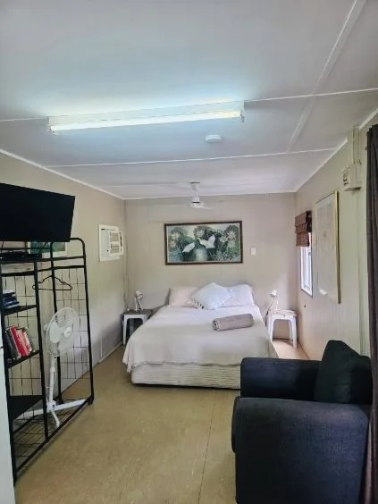 A simple bedroom with a bed, a black armchair, a small table, a flat-screen TV on a stand, and a painting on the wall.