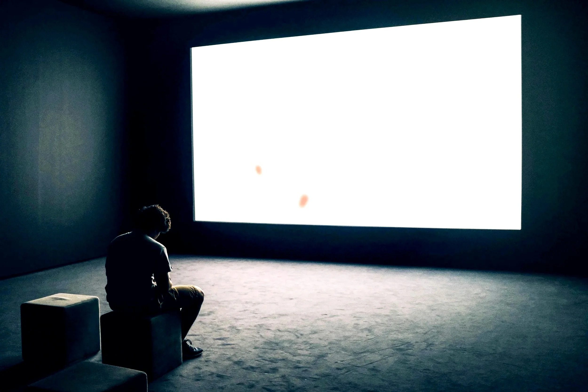 A person sitting on a small stool, alone in a dark room, watching a large bright screen.