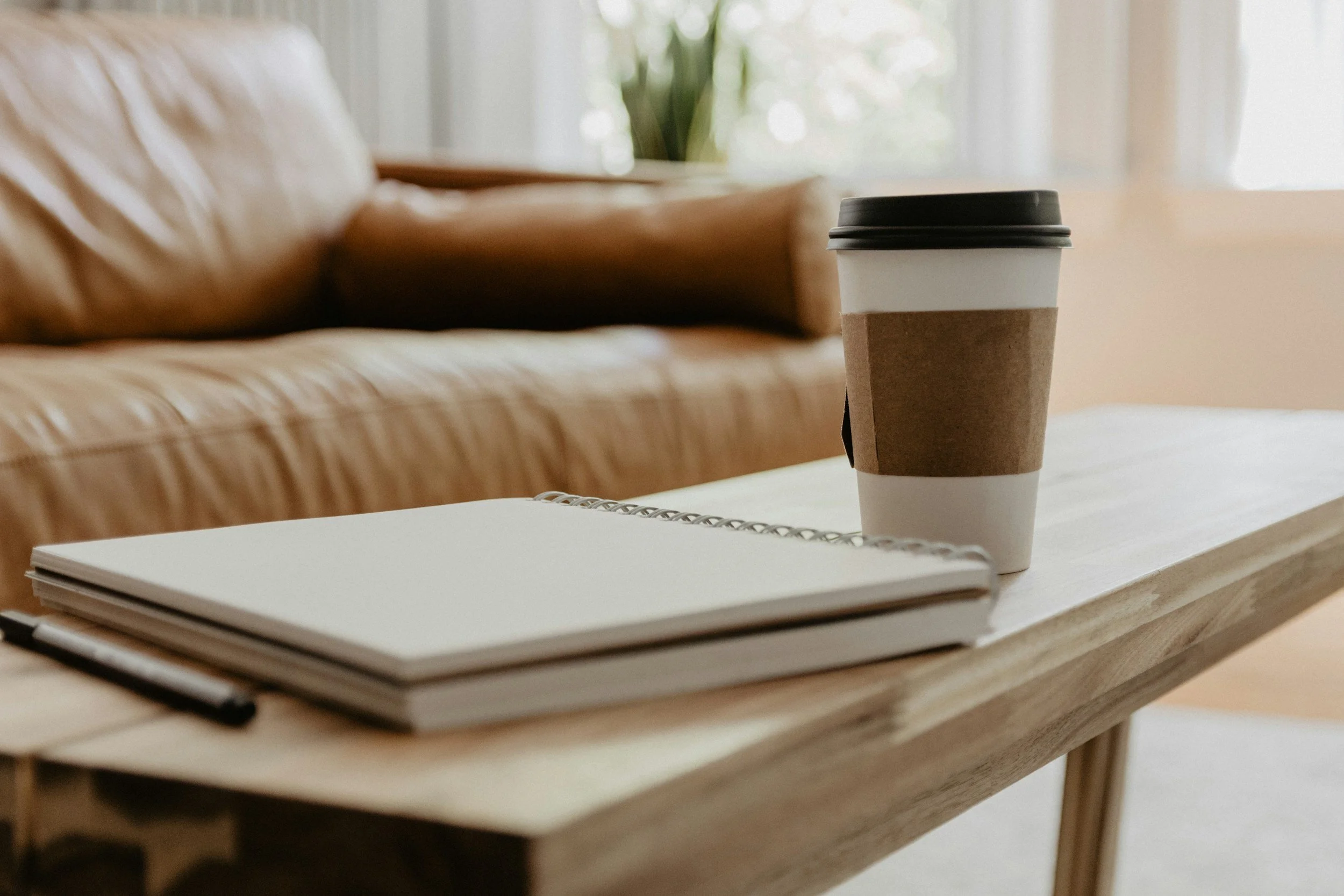 A wooden coffee table with a spiral notebook and pen, and a paper coffee cup with a black lid, in a cozy living room with a leather sofa and a window with curtains in the background.