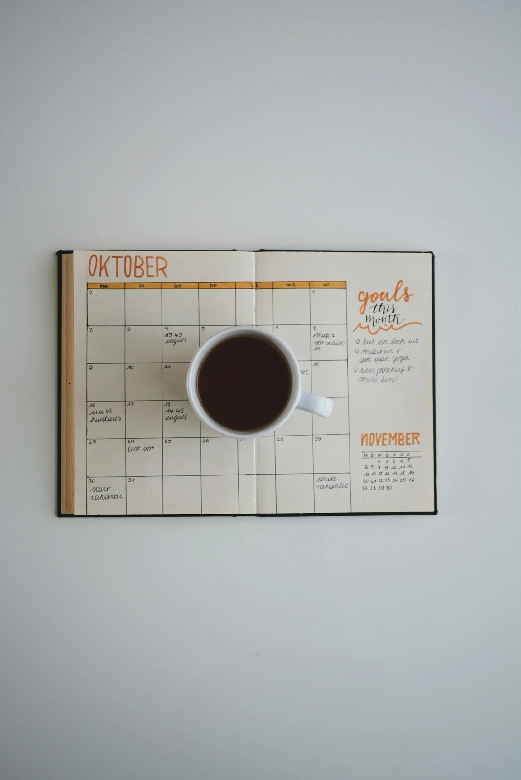 An open planner with handwritten notes for October and November, placed on a white surface, with a cup of coffee resting on the planner.