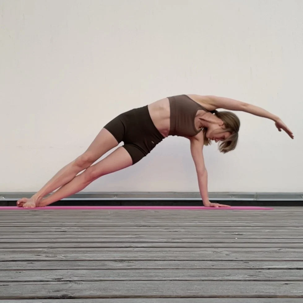 Woman performing a pilates side bend exercise on a pink mat on a wooden floor, wearing a brown sports bra and black shorts.
