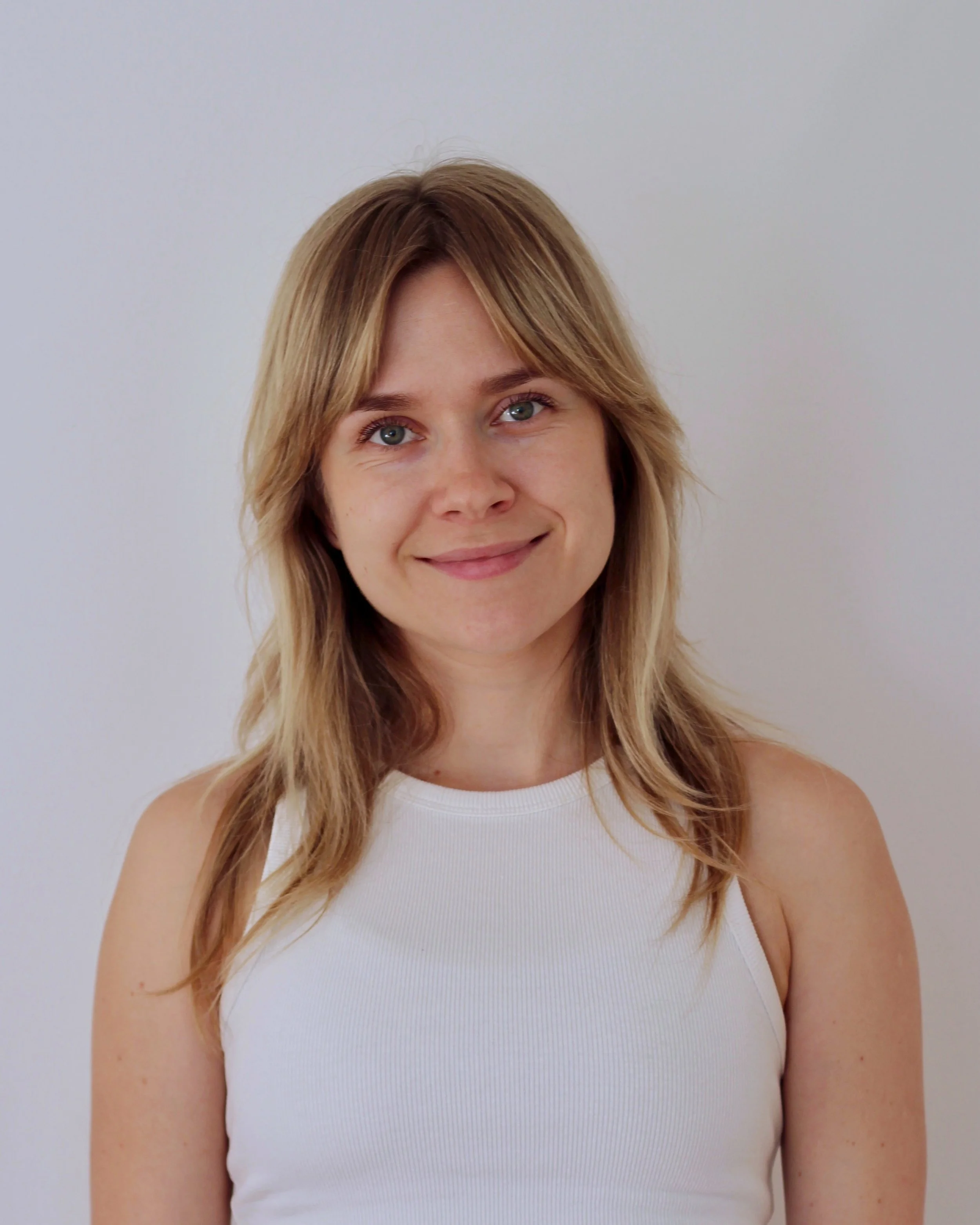 Young woman with shoulder-length light brown hair and blue eyes smiling at the camera, wearing a white sleeveless top, against a plain white background.