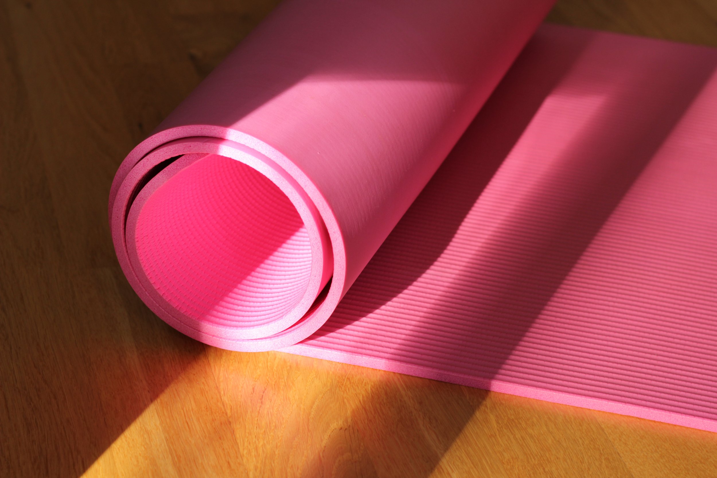 A pink yoga mat partially rolled on a wooden floor, with sunlight casting shadows.