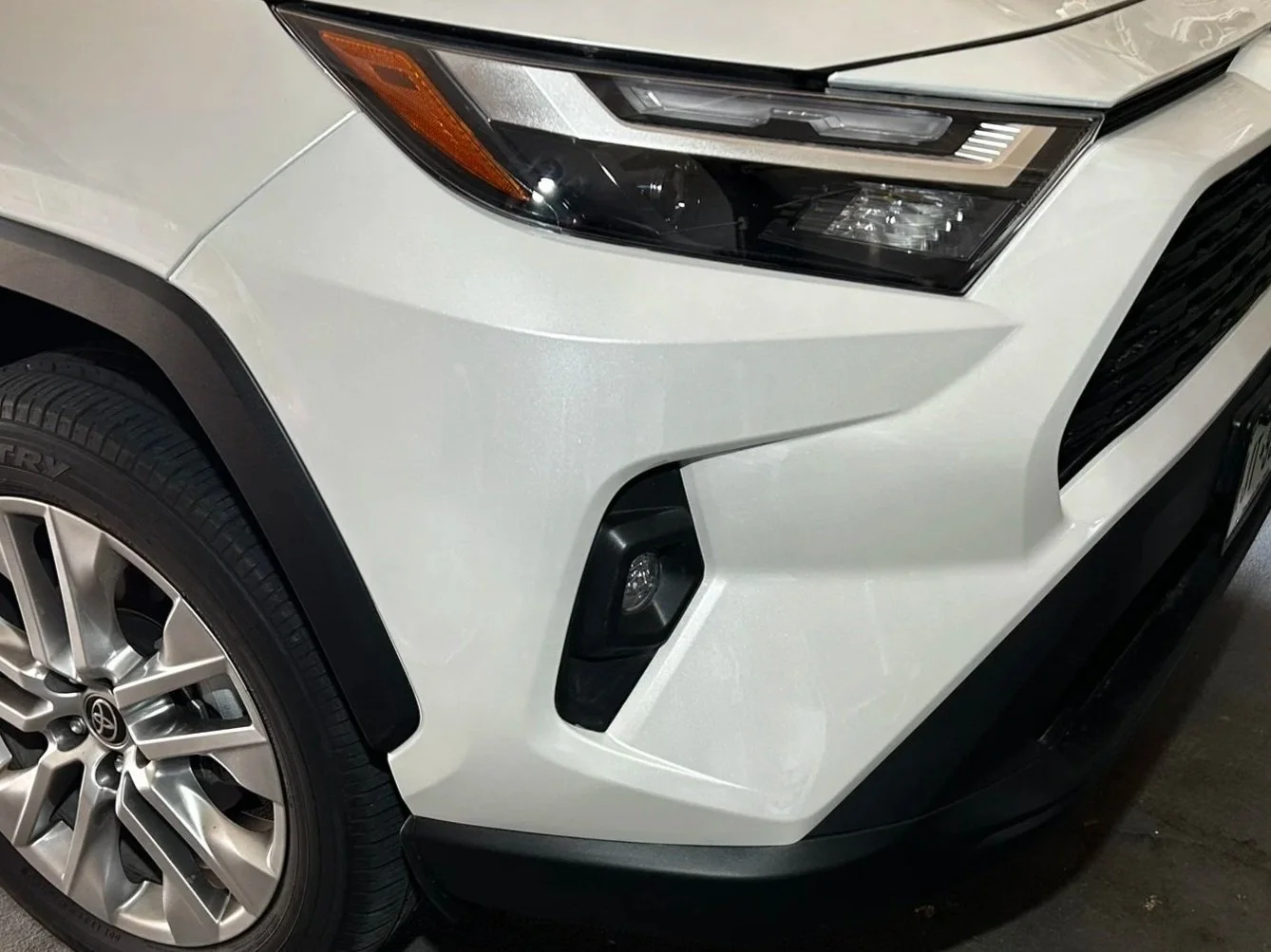 Close-up of the front right corner of a white car, showing the headlight, fog light, and wheel.