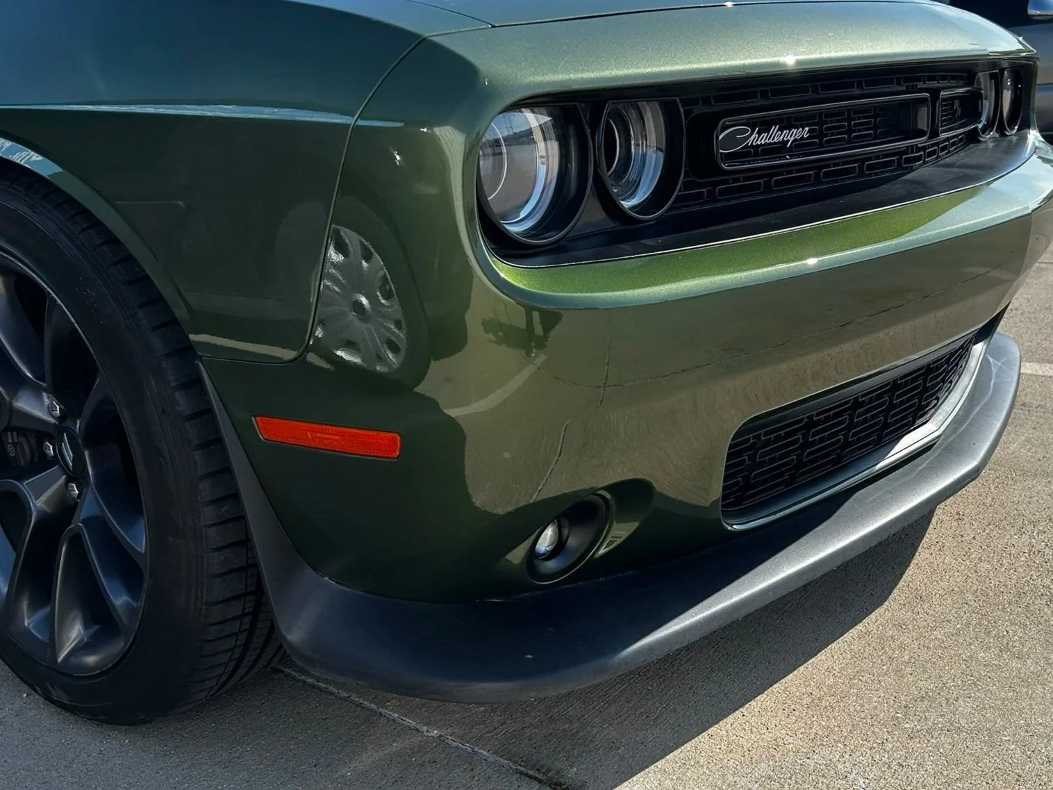 Front left side of a green Dodge Challenger car with black wheels, headlight, grille, and bumper.