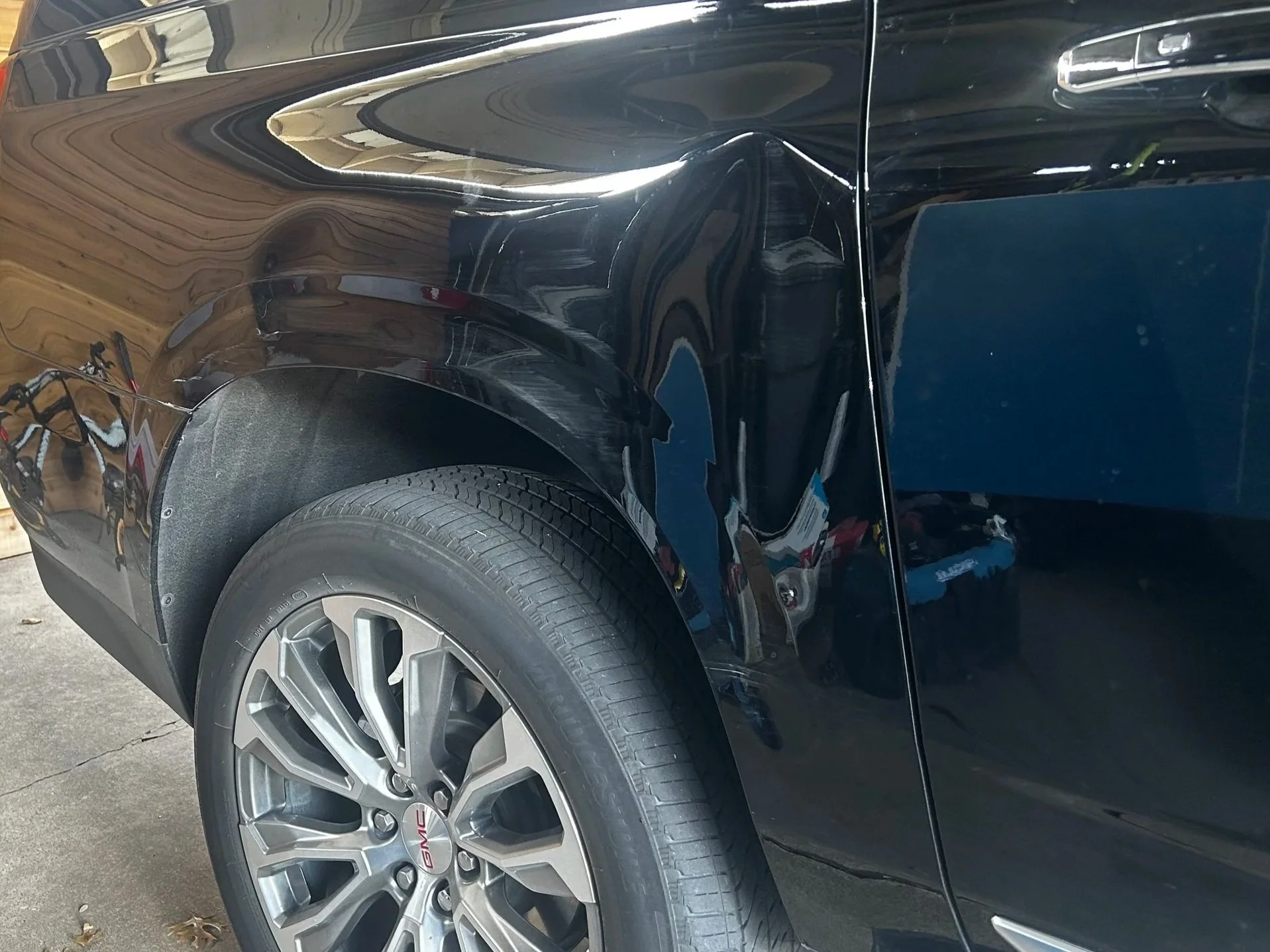 Close-up of the front right side of a black GMC vehicle, showing the wheel, tire, and part of the fender and door with a reflection of the surroundings.