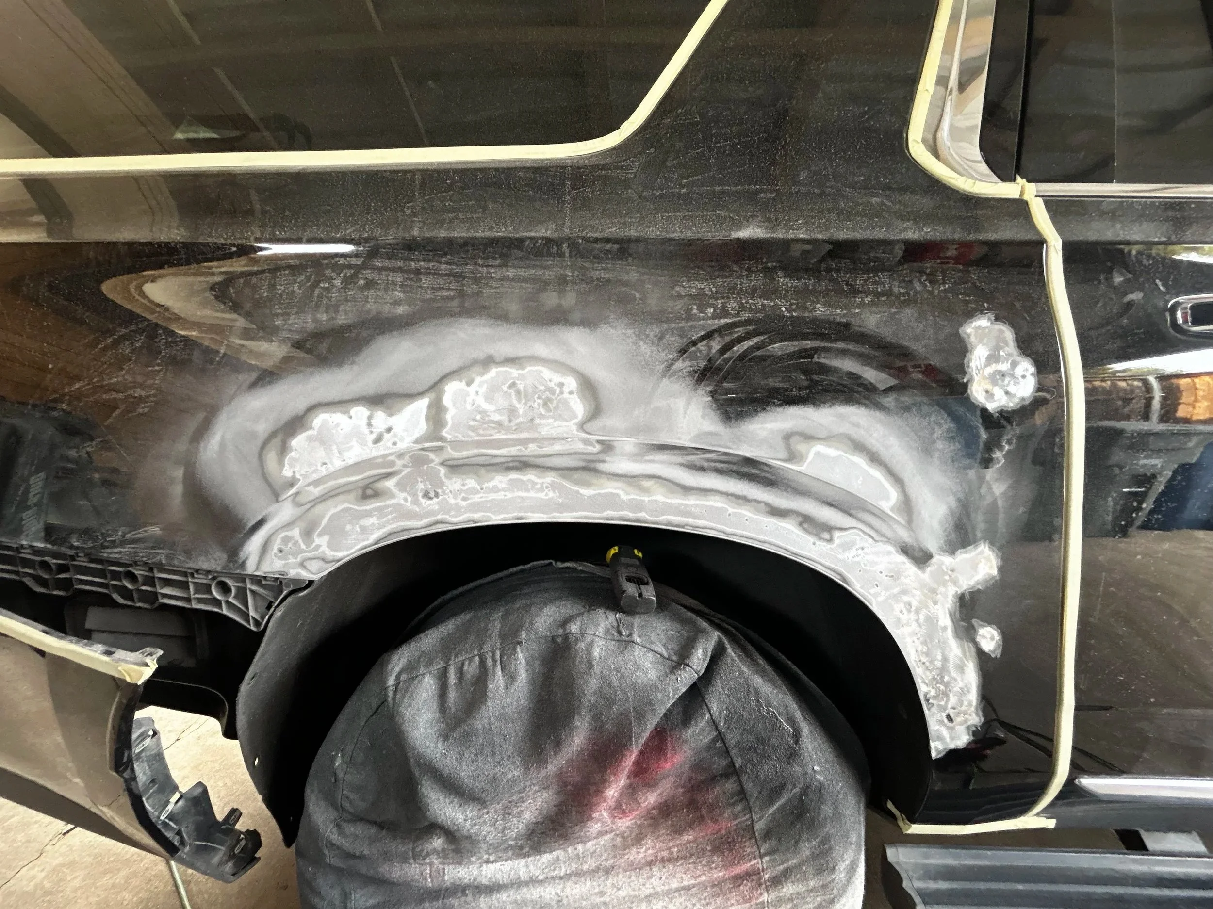 Close-up of a car's quarter panel under repair, showing sanding and primer application, with masking tape along the edges.