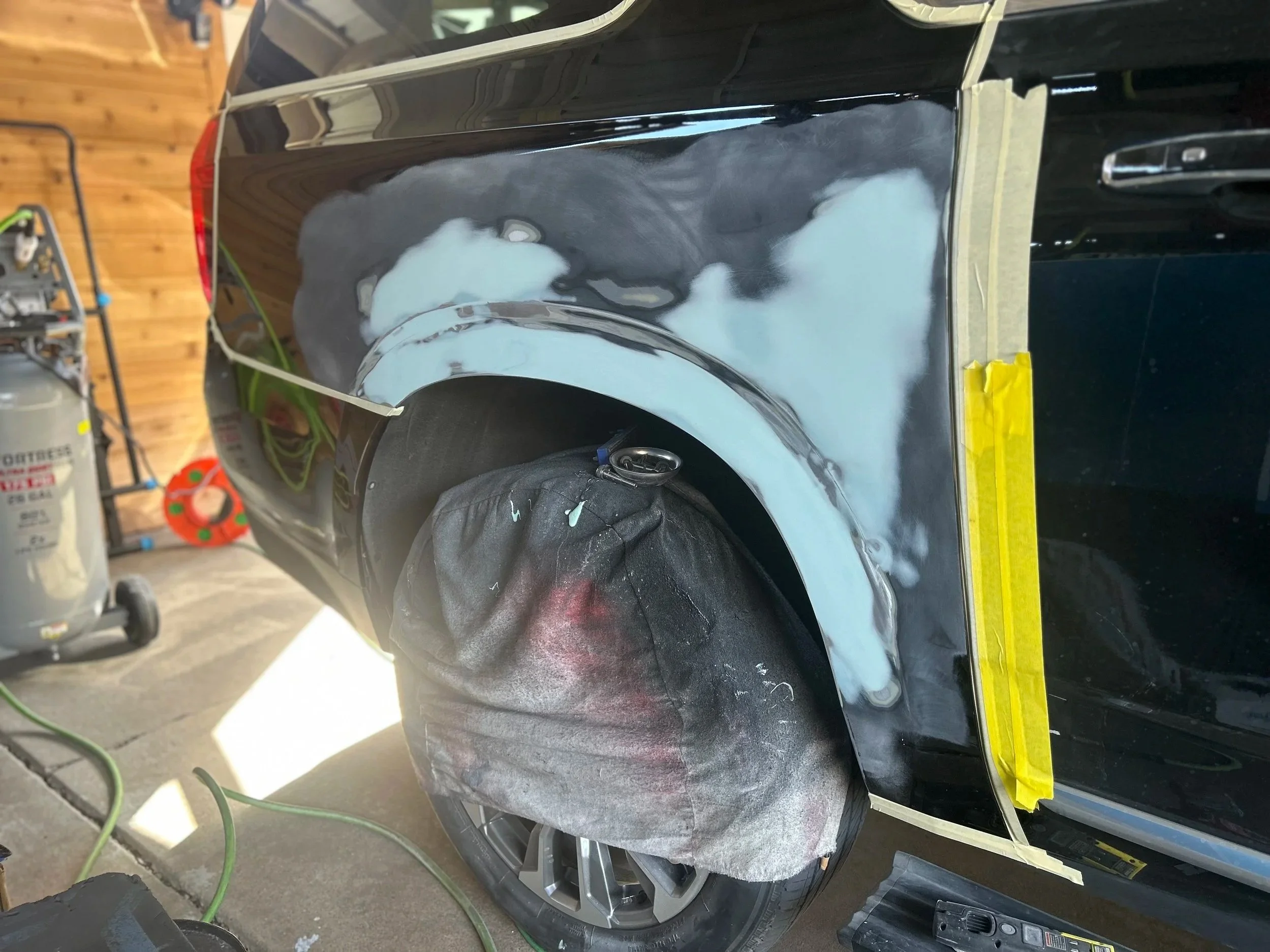 A black car in a garage being repaired or painted, with masking tape applied and patches of primer or filler on the body, surrounding the wheel.
