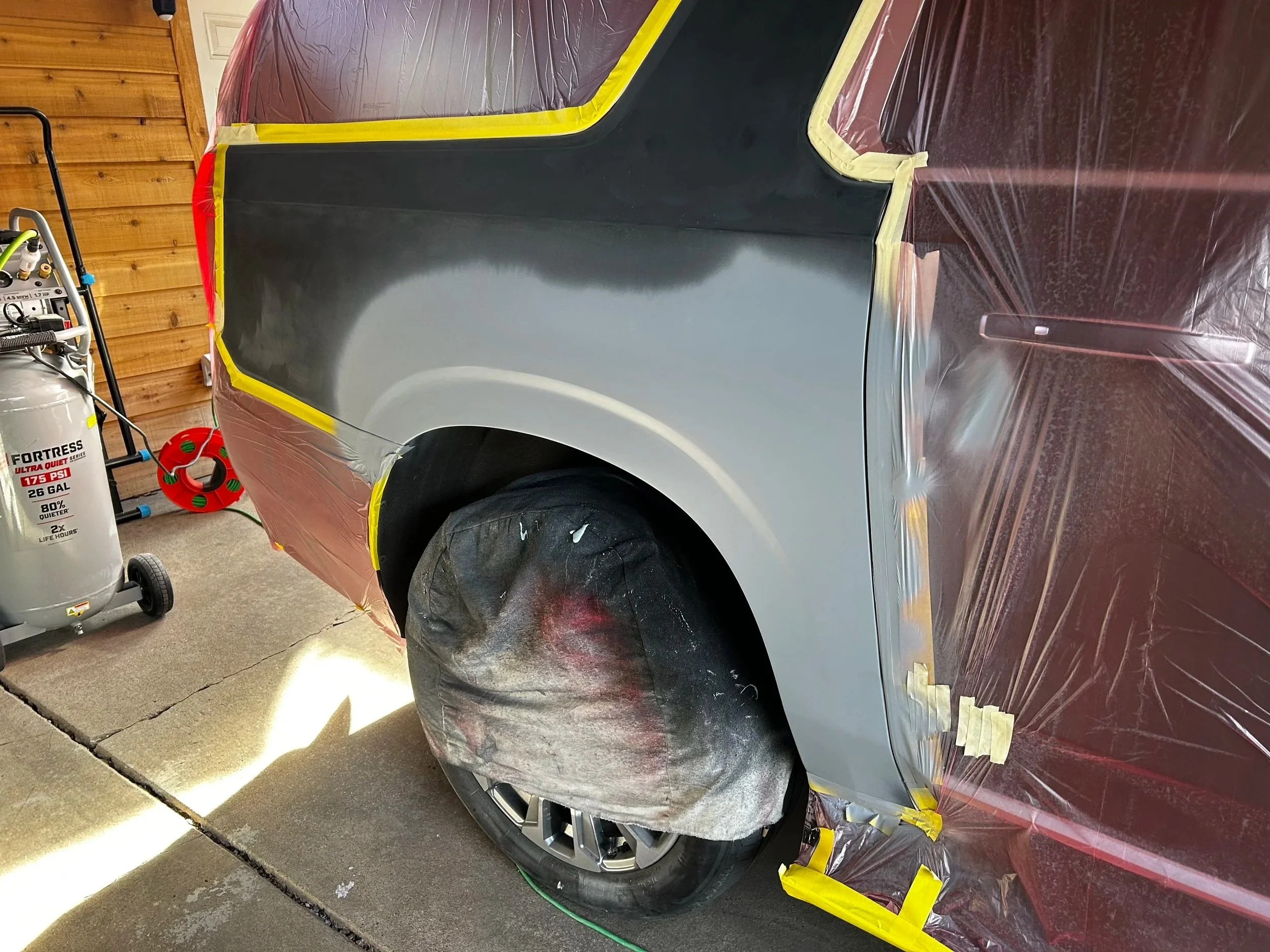Car being painted with gray primer, protected with plastic sheets and masking tape.