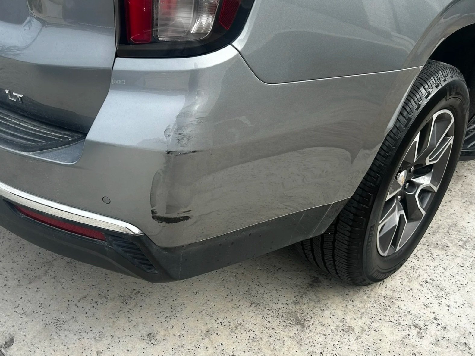 The rear right side of a gray SUV shows scratches and a dent on the bumper and wheel arch.