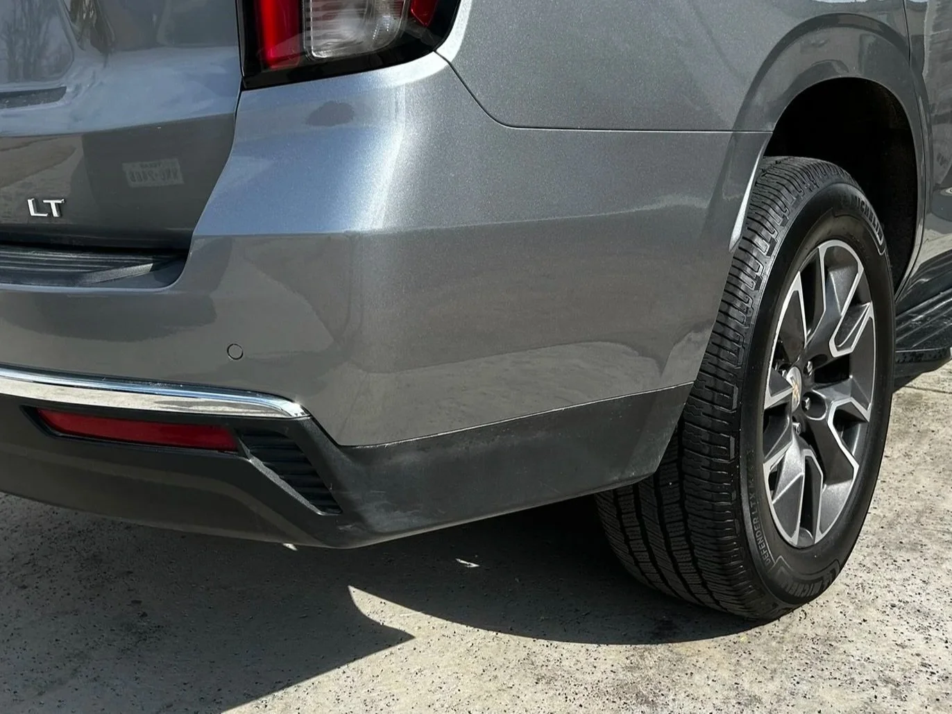 Close-up of the rear right side of a gray car, showing the wheel, tire, rear bumper, and part of the tail light.