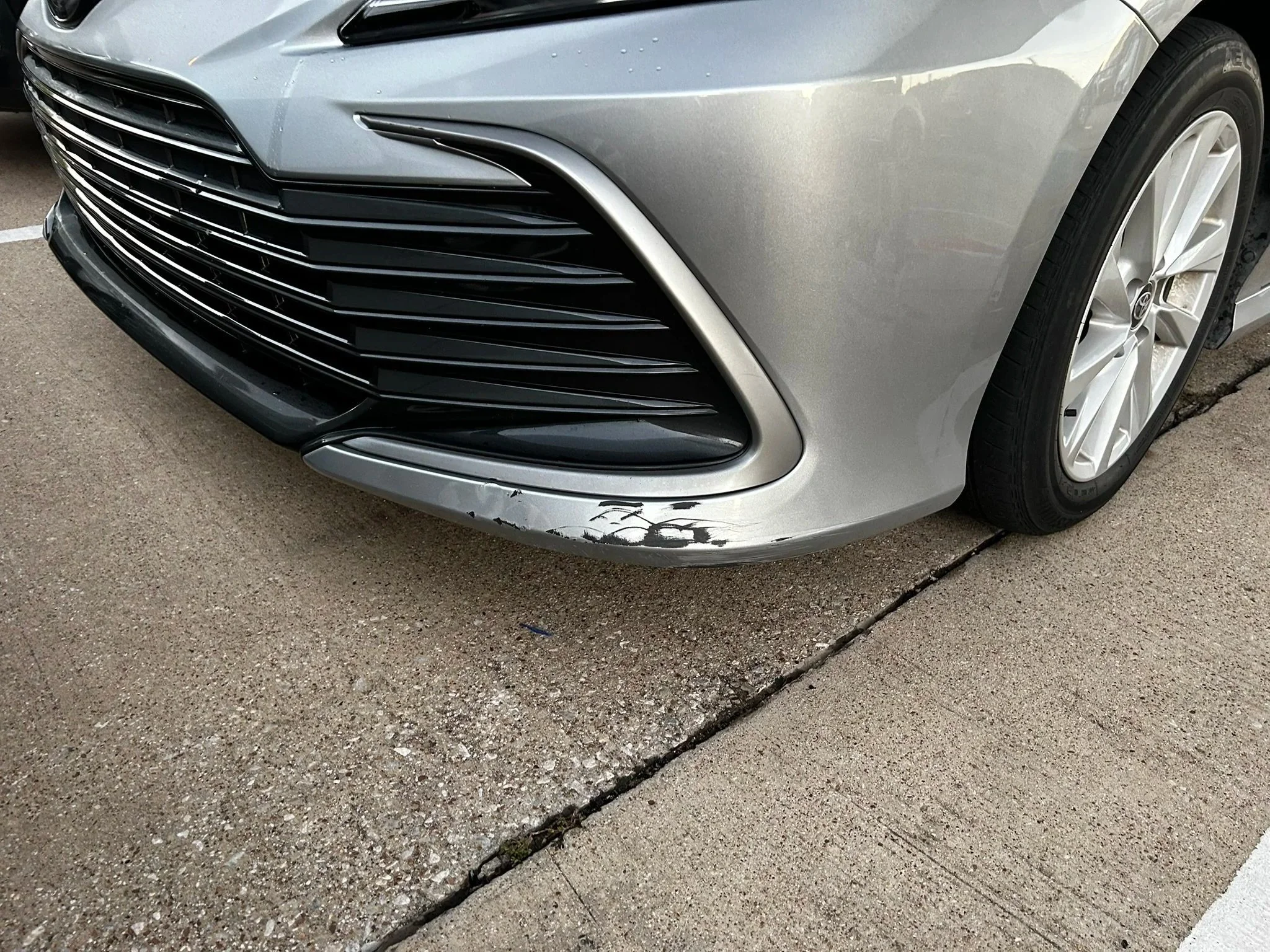 Front of a silver car with black grille and minor scratch on the bumper, parked on concrete pavement.