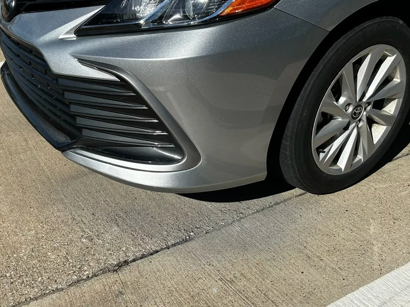 Close-up of the front right corner of a silver sedan car showing the headlight, grille, wheel, and part of the pavement.
