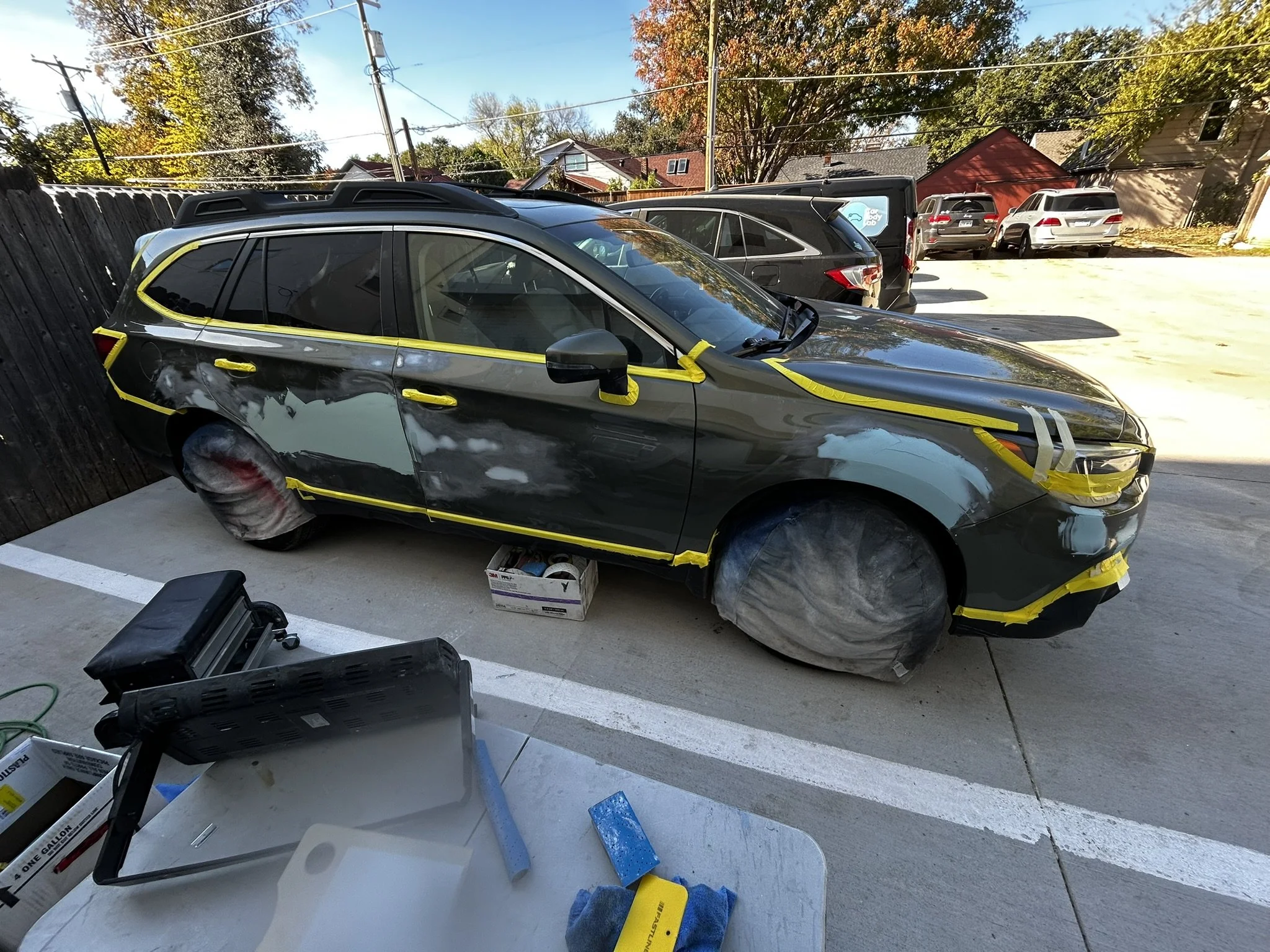 A black station wagon car being painted or repaired outdoors. The car is masked with tape and paper, with some areas in primer or paint. The hood and front of the car are masked, and the rear wheel is covered with protective paper. There are tools an