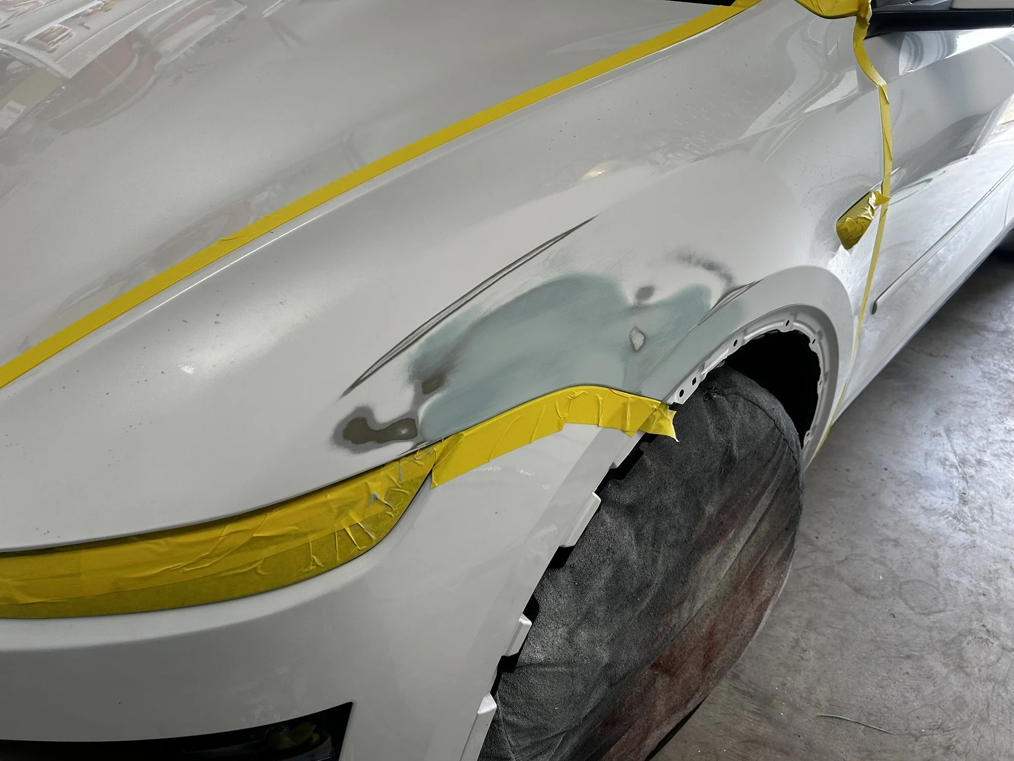 A white car undergoing body repair with visible sanding and primer work, yellow masking tape applied around the fender and headlight area, and a black tire covered with a protective cloth.