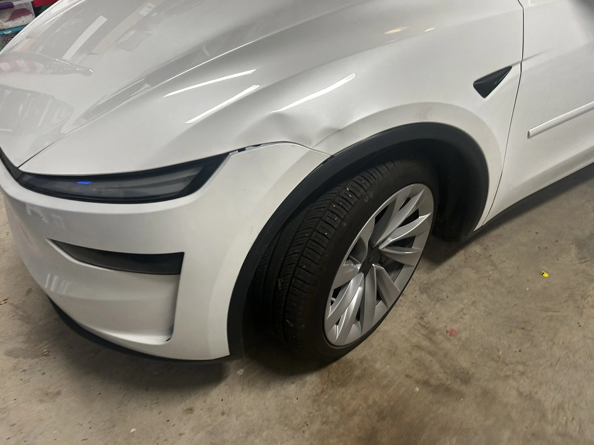 Close-up of the front left side of a white Tesla Model 3, showing the headlight, front wheel, and part of the front bumper in a garage.