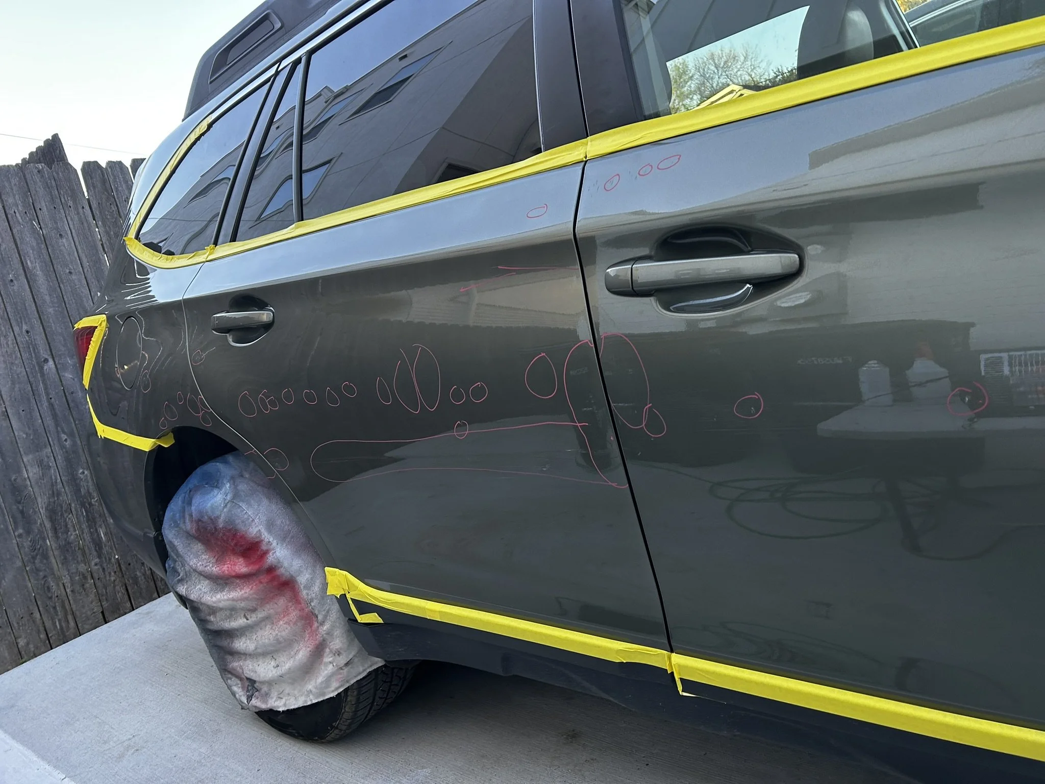 A gray car with yellow masking tape around the windows, doors, and wheel arch. The car has red circle markings and spray painted circles on the body, likely for a repair or painting process. The car is parked outside near a wooden fence.