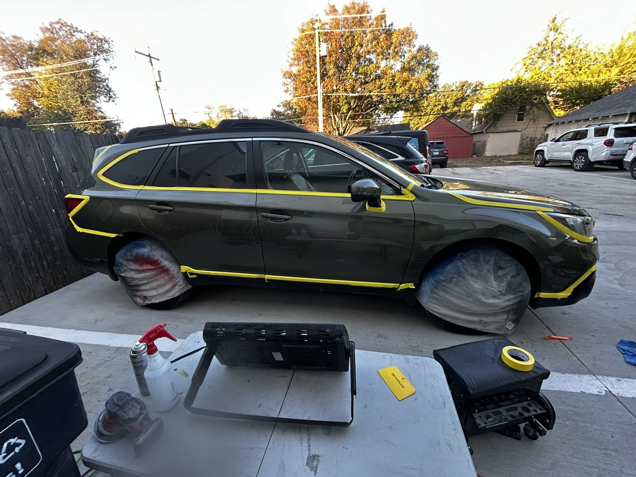 A black SUV is parked outdoors on a concrete surface with yellow masking tape on the windows and wheels covered with protective material, likely for painting or detailing. There are tools, spray bottles, and a laptop on a nearby table.