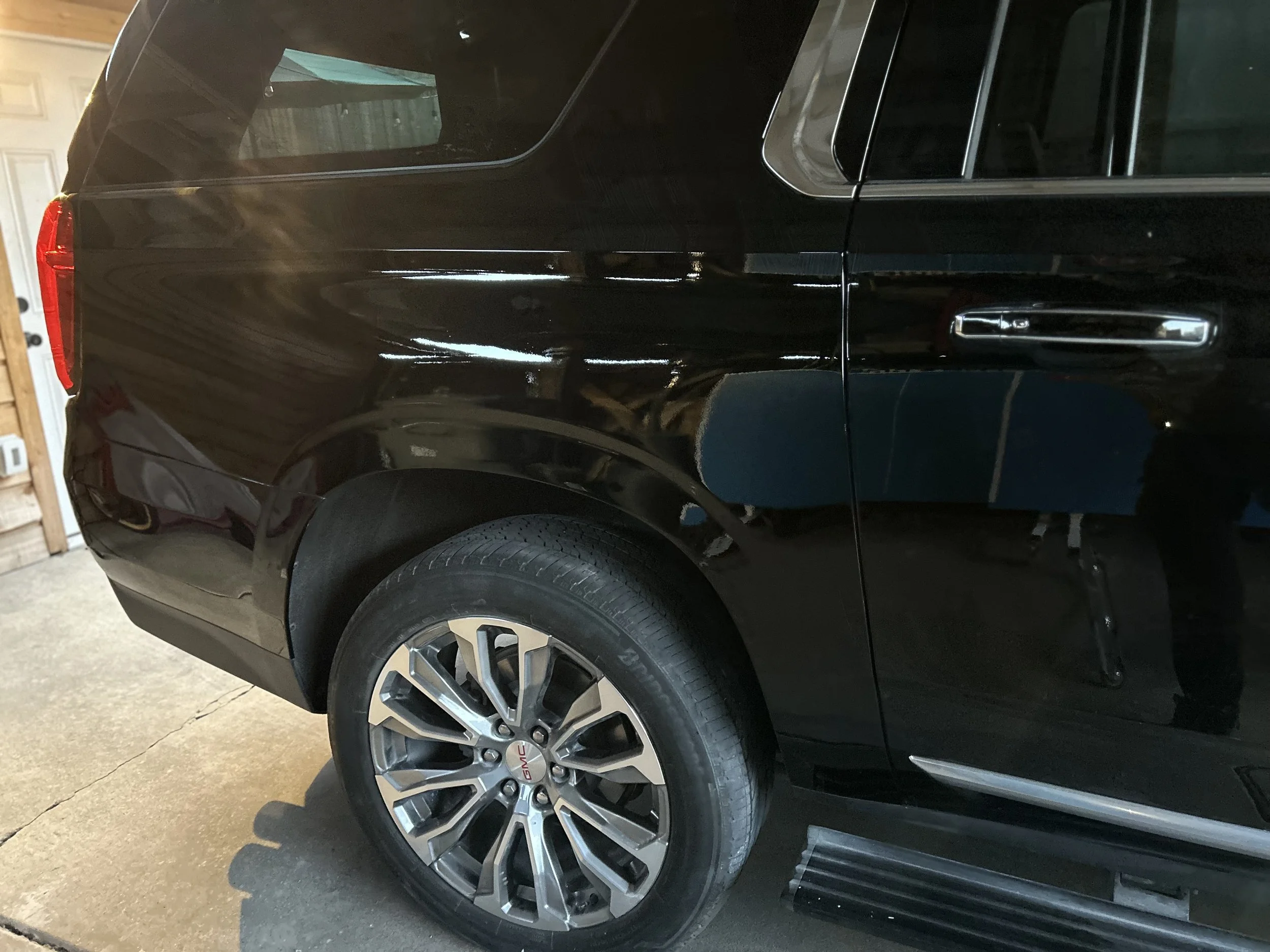 Close-up of the rear side of a shiny black GMC SUV parked inside a garage, featuring a wheel with a multi-spoke rim and part of the garage door in the background.
