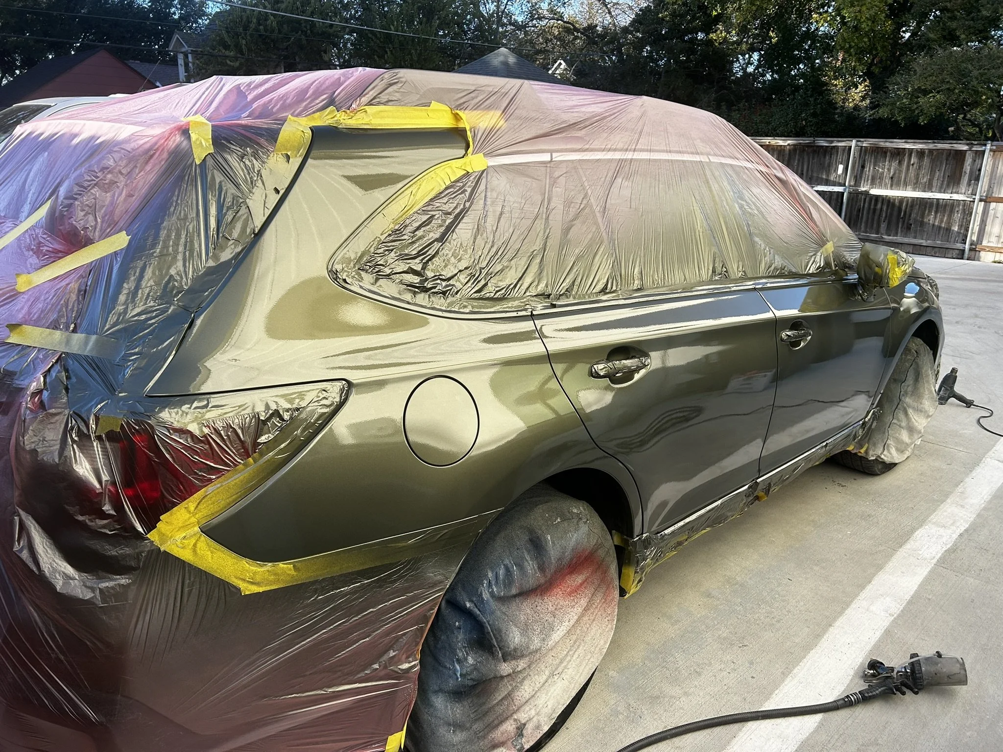 A car in the process of being painted, wrapped in protective plastic sheeting and masking tape, with sanding tools nearby.