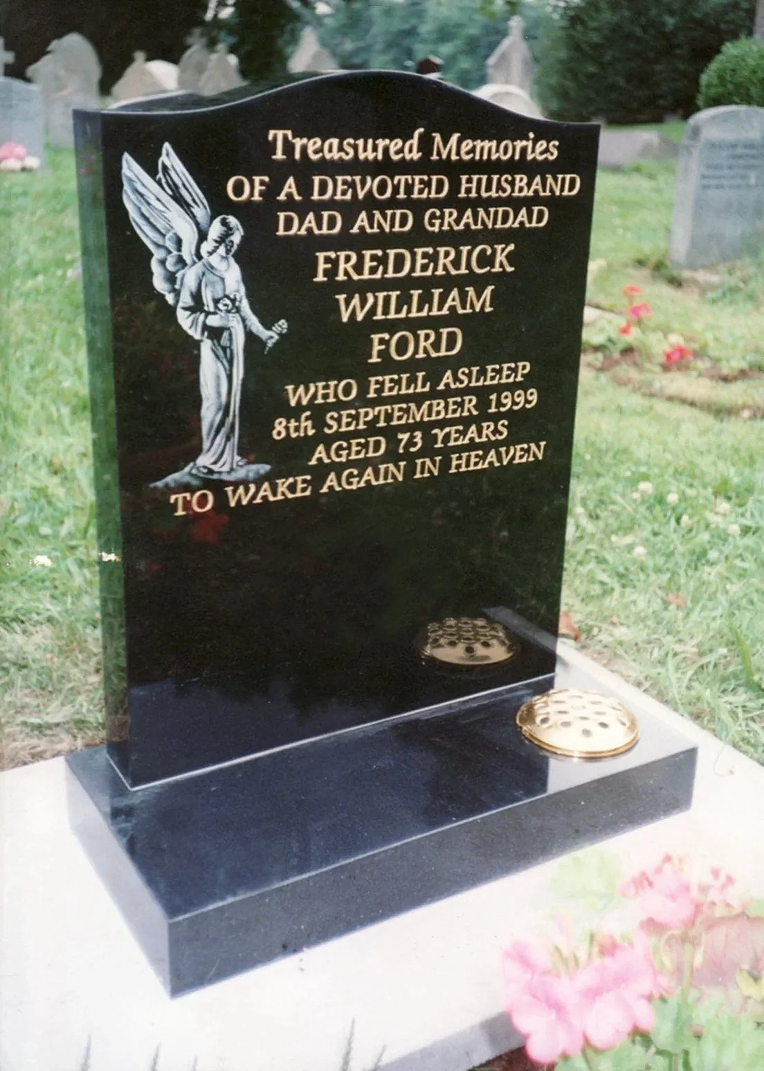 Black polished headstone with engraved angel and memorial text in a cemetery, flowers nearby.
