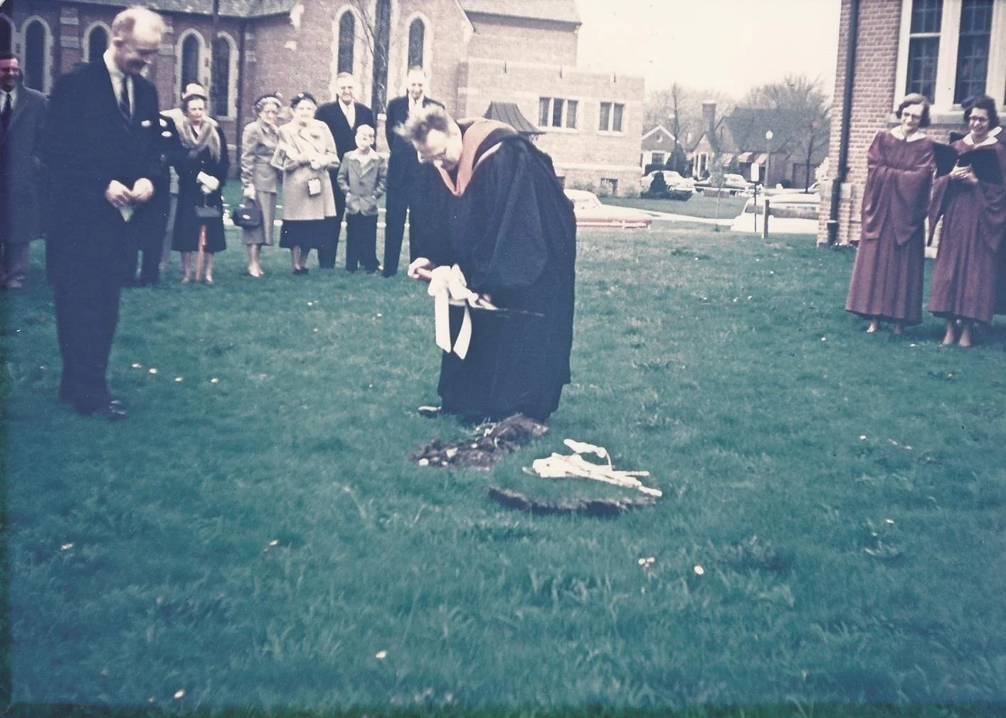 A woman dressed in a black academic gown and hood is performing a ceremony, possibly planting a tree, on a rainy day outside a brick building. Several people in formal attire, including men and women, are gathered around watching, some holding umbrellas, with houses and parked cars in the background.