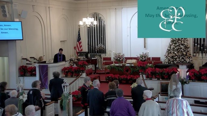 People gather inside a church decorated for Christmas, with a Christmas tree, poinsettias, and an organ in the background. Some individuals are standing, while others are seated near the front. A pastor is at the pulpit, and a screen displays lyrics 