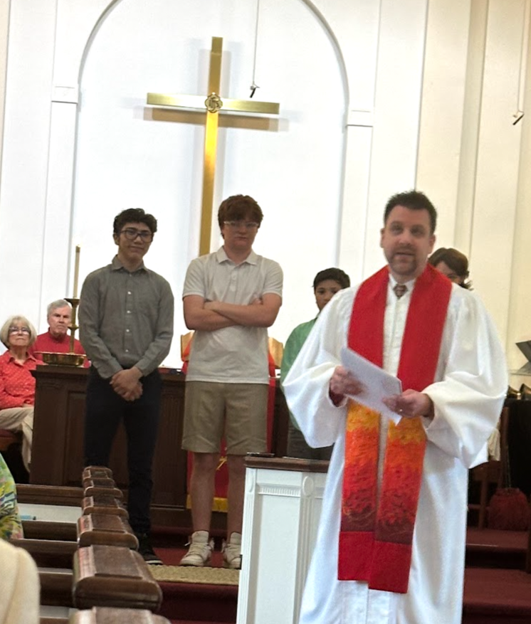 A church service with a priest holding papers, standing in front of a congregation, with a cross and an altar in the background. Three young men stand behind him.