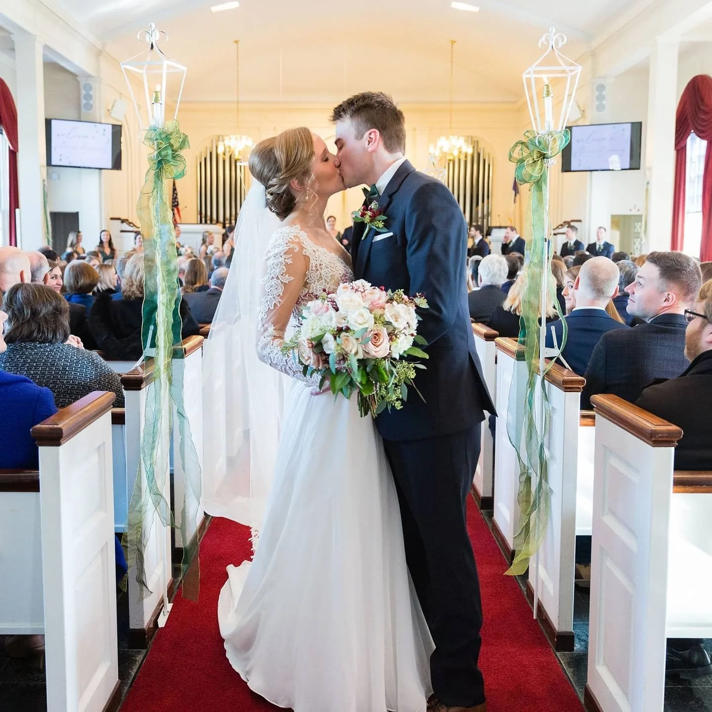 A bride and groom share a kiss during their wedding ceremony in a church, with guests seated in pews in the background.