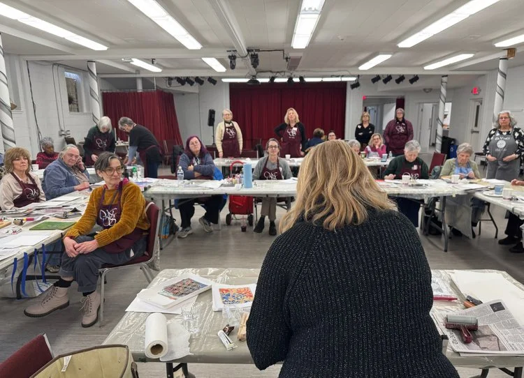 A group of women gathered in a room with tables, some standing and some seated, participating in a workshop or meeting. The room has a red curtain on the stage area at the back and various papers and materials on the tables.