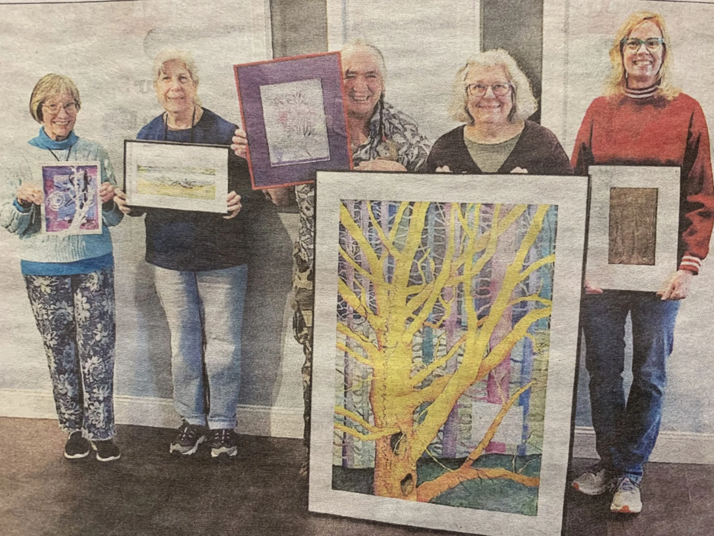 Six women standing side by side, each holding framed artwork, with a colorful painting of a tree in the foreground.